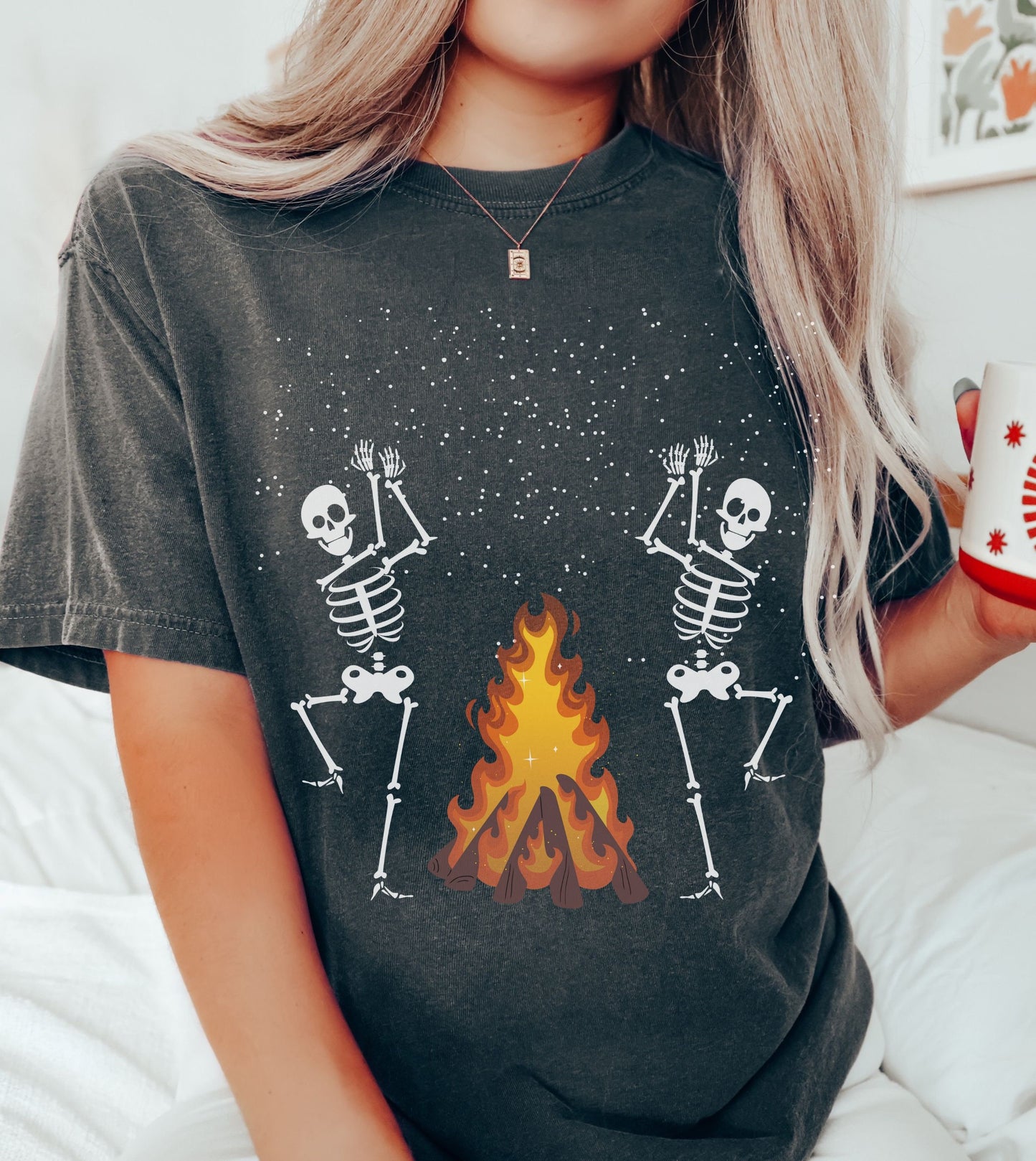 A woman wearing a cute pepper colored shirt with two white skeletons dancing and clapping hands around a roaring bonfire.