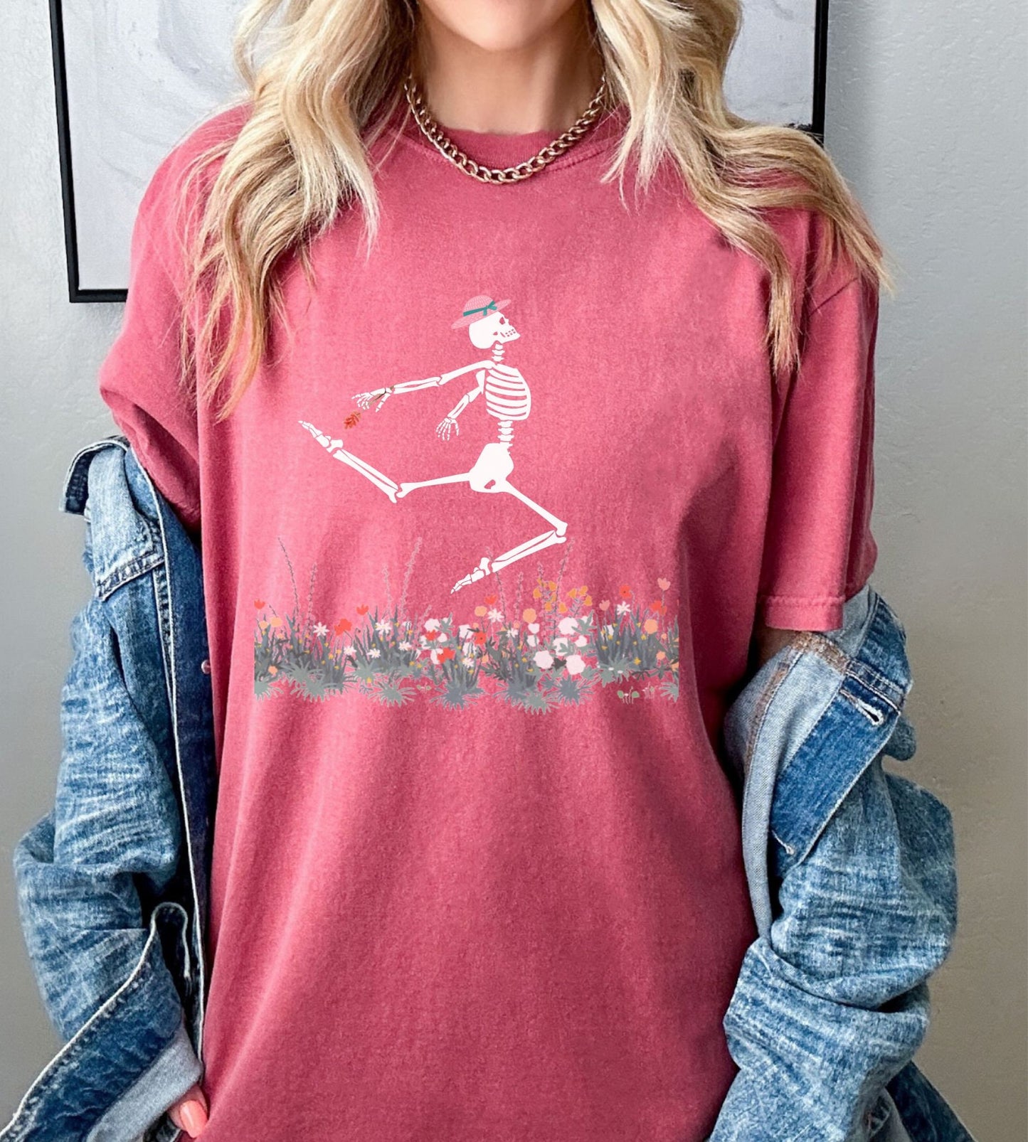 A woman wearing a cute maroon colored shirt with a white female skeleton in a pink bonnet holding a red flower dancing in the air above a meadow of flowers.