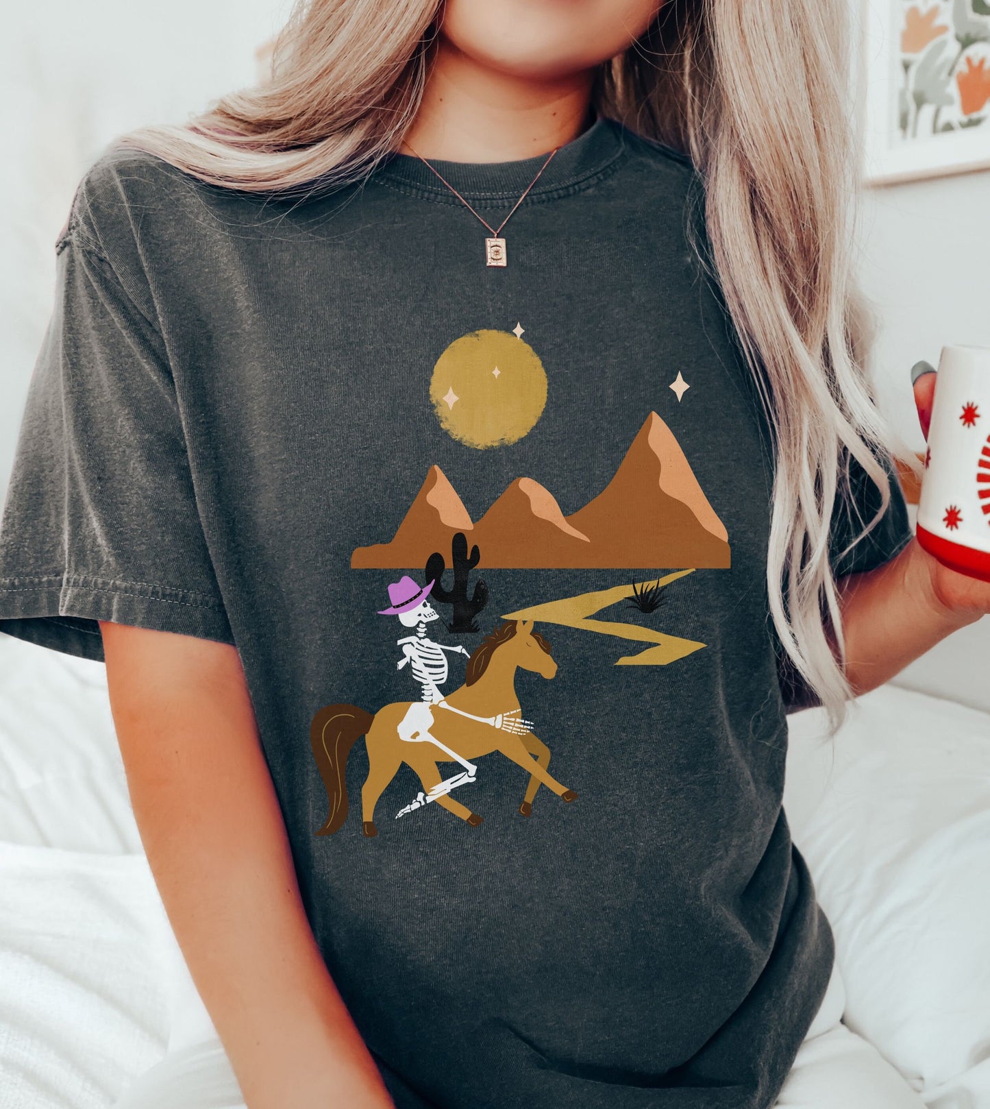 A woman wearing a vintage, pepper colored shirt with a skeleton cowgirl wearing a pink cowboy hat riding a brown horse through the eset at night, cacti and three mountains in the background with a full moon and stars in the sky.