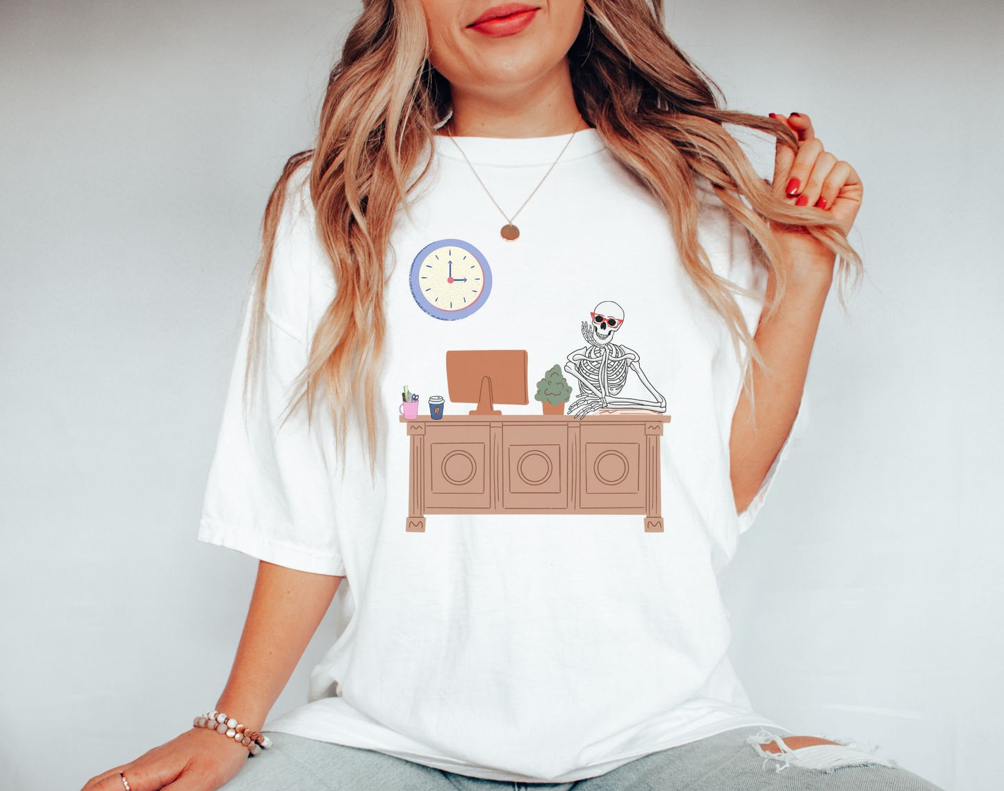 A woman wearing a cute white colored shirt with a female skeleton wearing red glasses sitting at a desk leaning her head on one hand, a plant and desktop computer to her right with a pen holder and coffee to the right.
