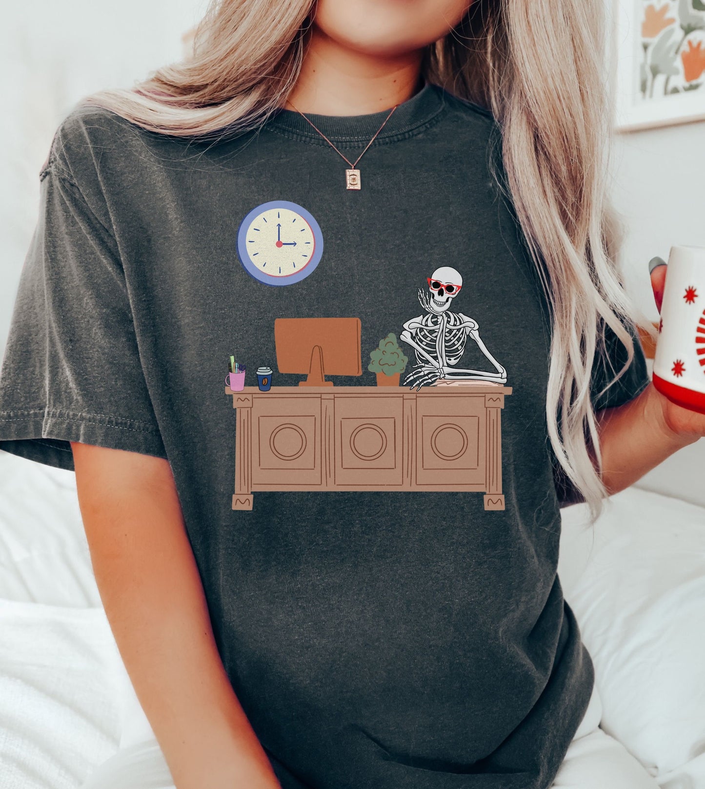 A woman wearing a cute black colored shirt with a female skeleton wearing red glasses sitting at a desk leaning her head on one hand, a plant and desktop computer to her right with a pen holder and coffee to the right.