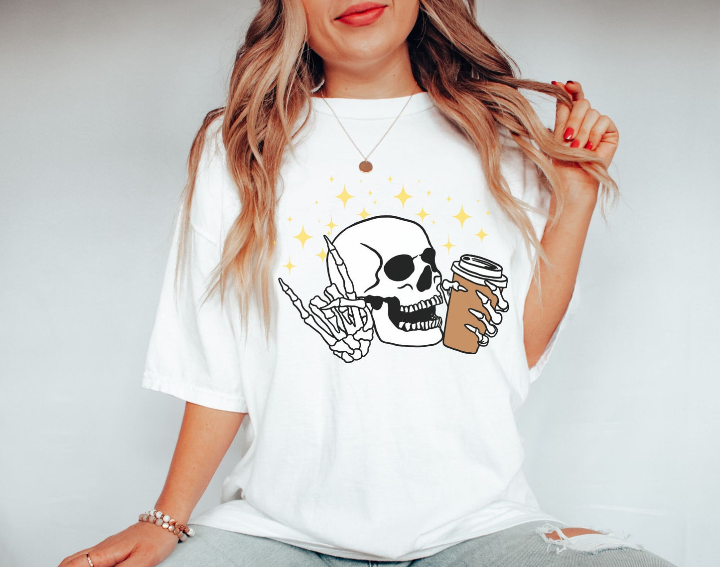 A woman wearing a cute white colored shirt, a skeleton skull holding a coffee in the left hand and holding up the rock on hand sign with the right hand with twinkly stars in the background.