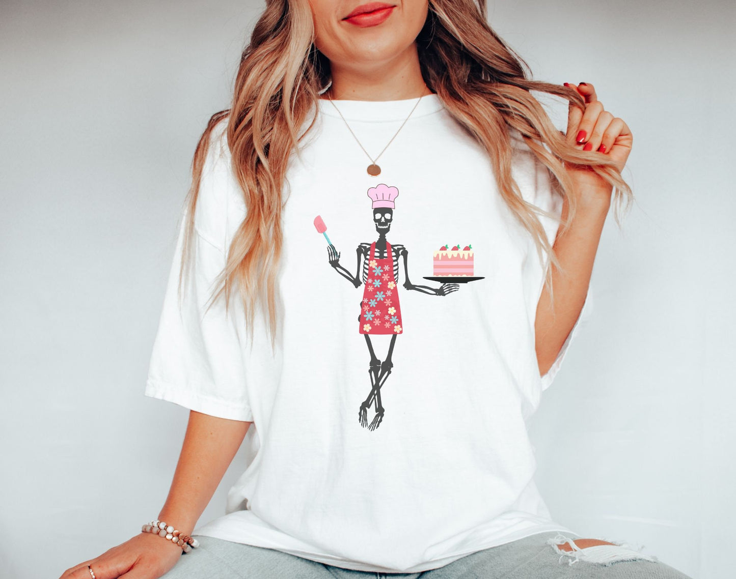A woman wearing a cute white colored shirt with a female skeleton wearing a pink chef’s hat standing cross legged in a red apron adorned with flowers holding a cake on a plate in her left hand and a spatula in her right hand.