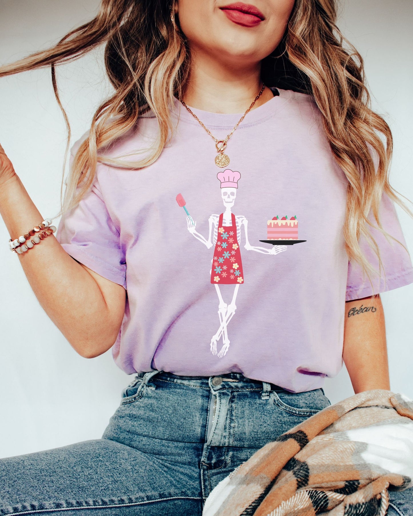 A woman wearing a cute light purple colored shirt with a female skeleton wearing a pink chef’s hat standing cross legged in a red apron adorned with flowers holding a cake on a plate in her left hand and a spatula in her right hand.