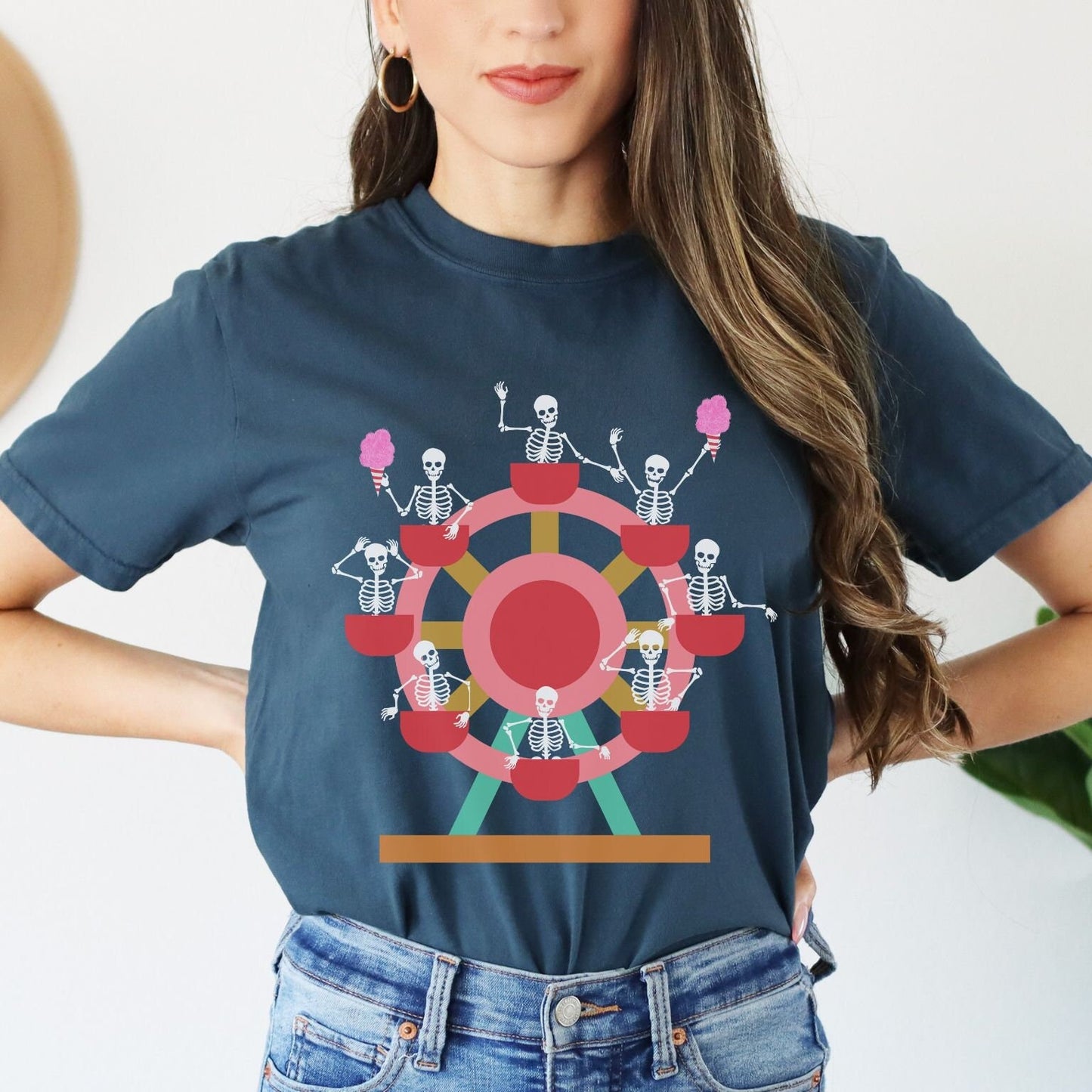 A woman wearing a cute midnight colored shirt with a ferris wheel filled with black skeletons waving and holding cotton candy as they go around the ride.