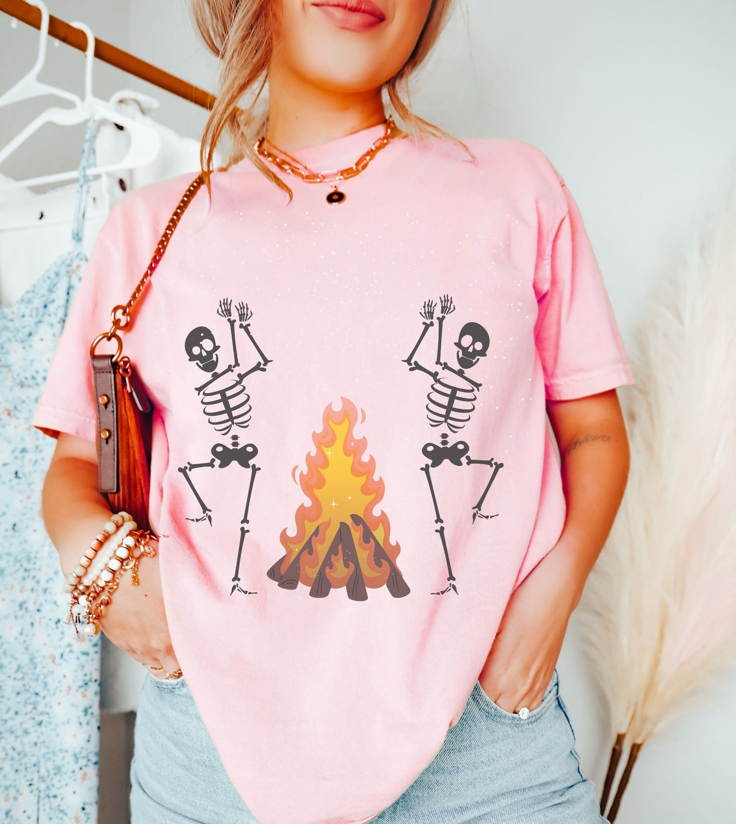 A woman wearing a cute light pink colored shirt with two white skeletons dancing and clapping hands around a roaring bonfire.