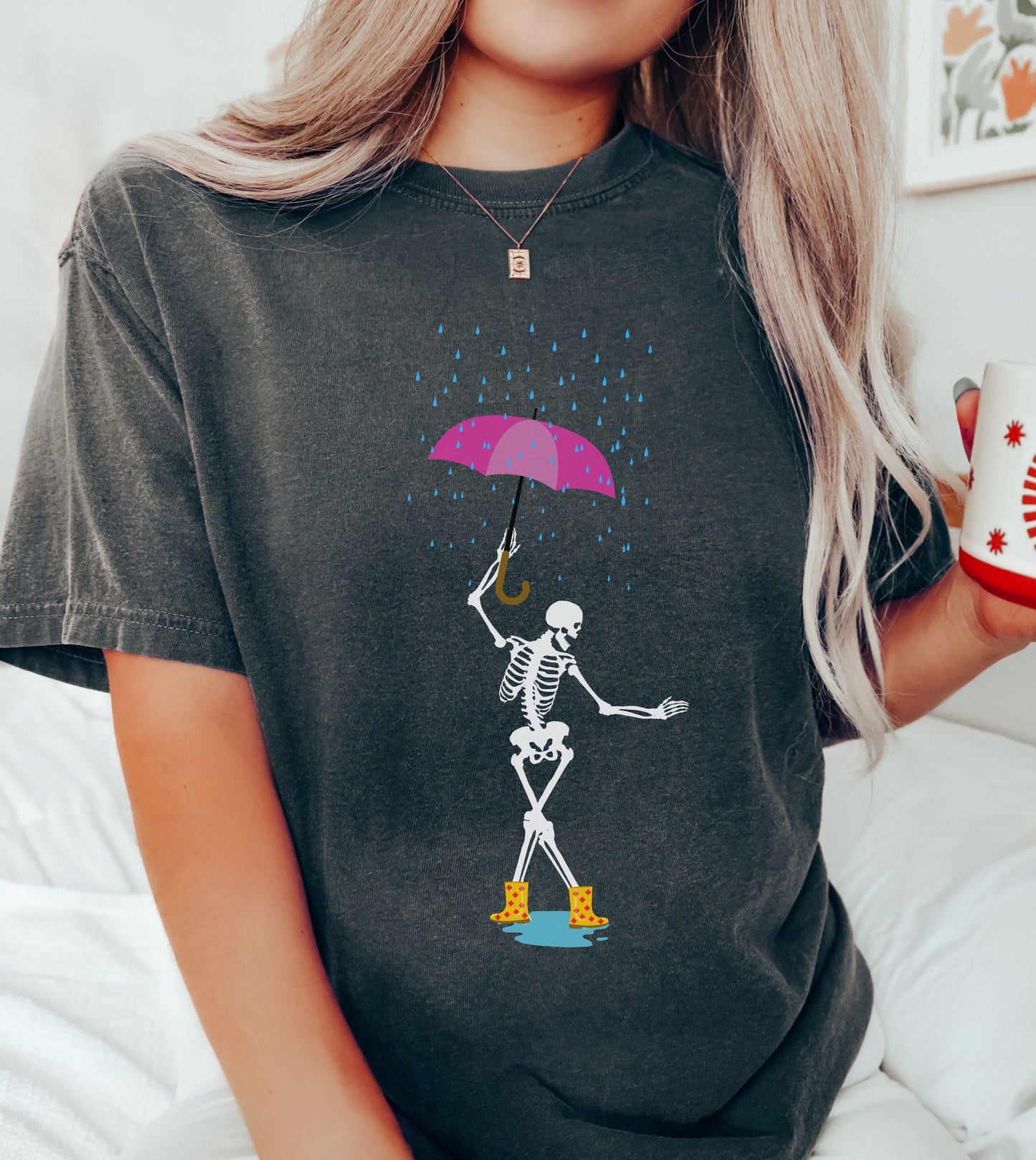 A woman wearing a cute pepper colored shirt with a female skeleton dancing in the rain with a pink and purple umbrella and wearing yellow with red polka dot rain boots.