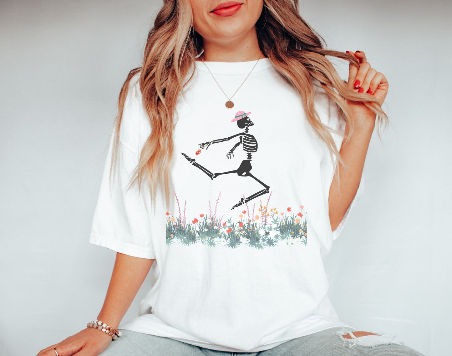 A woman wearing a cute white colored shirt with a white female skeleton in a pink bonnet holding a red flower dancing in the air above a meadow of flowers.