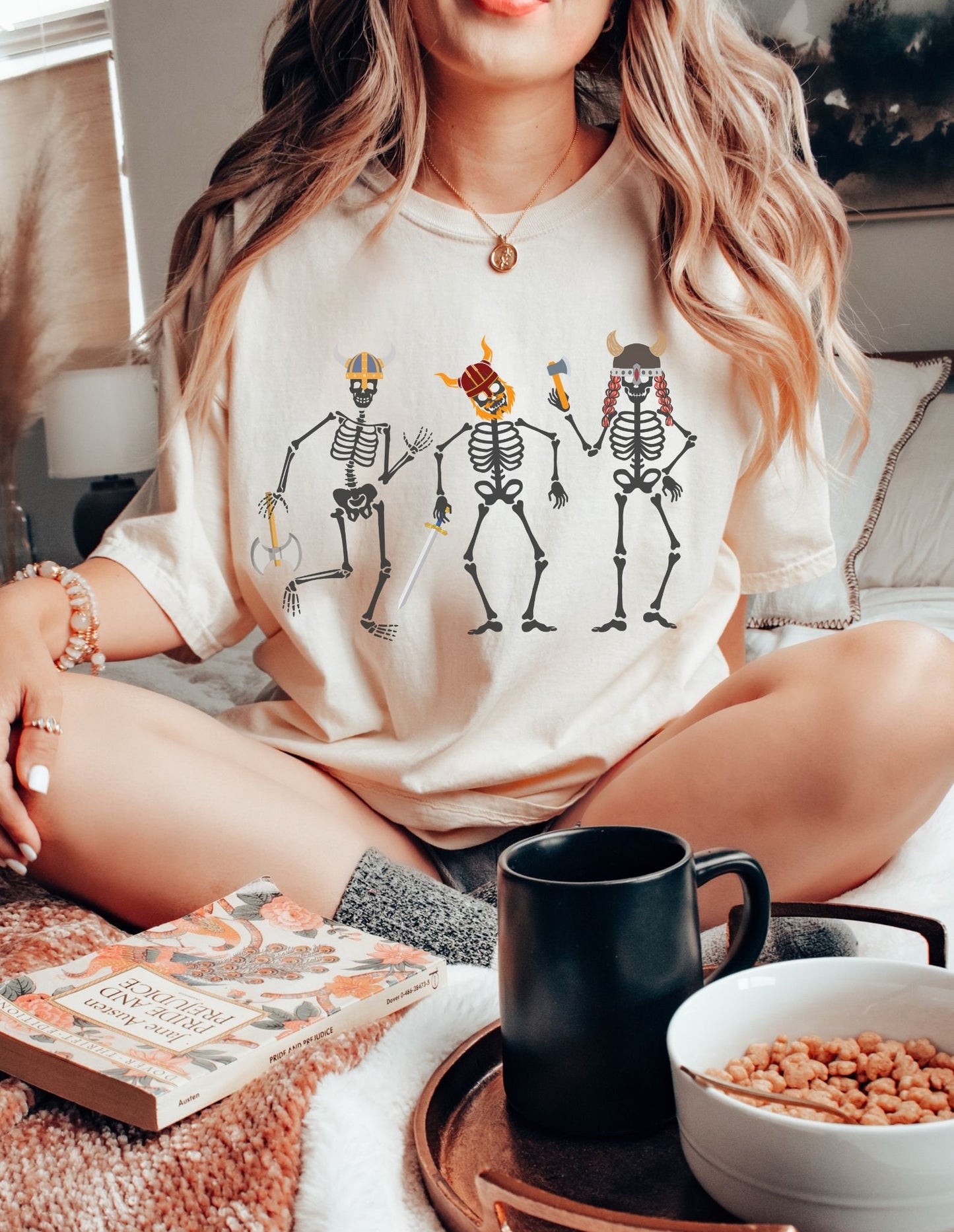 A woman wearing a vintage, ivory colored shirt with three skeletons wearing horned helmets dressed as vikings. One is holding a battle axe, another has a red beard and a sword, and the third has long red hair and is wielding a hatchet.