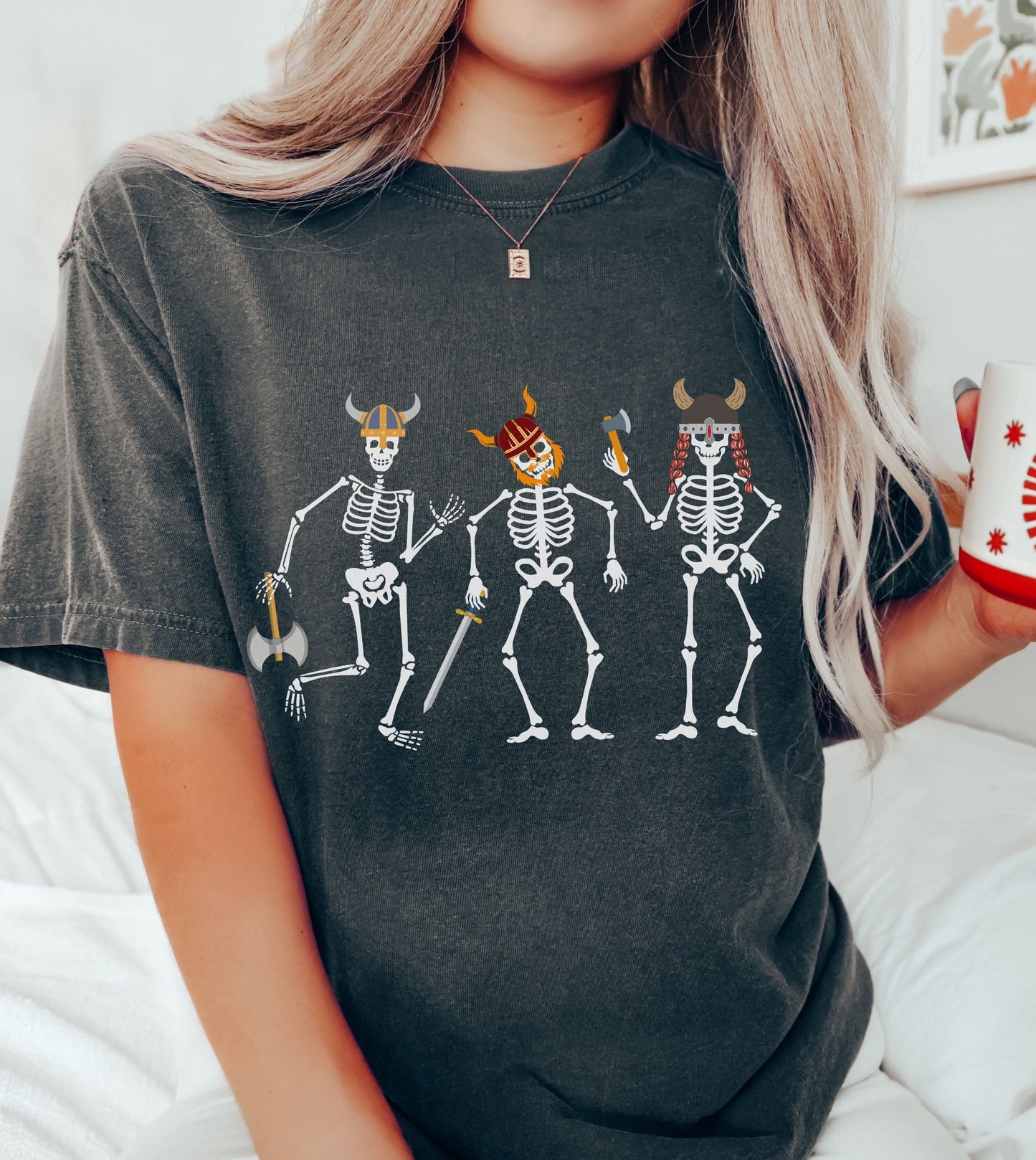 A woman wearing a vintage, pepper colored shirt with three skeletons wearing horned helmets dressed as vikings. One is holding a battle axe, another has a red beard and a sword, and the third has long red hair and is wielding a hatchet.