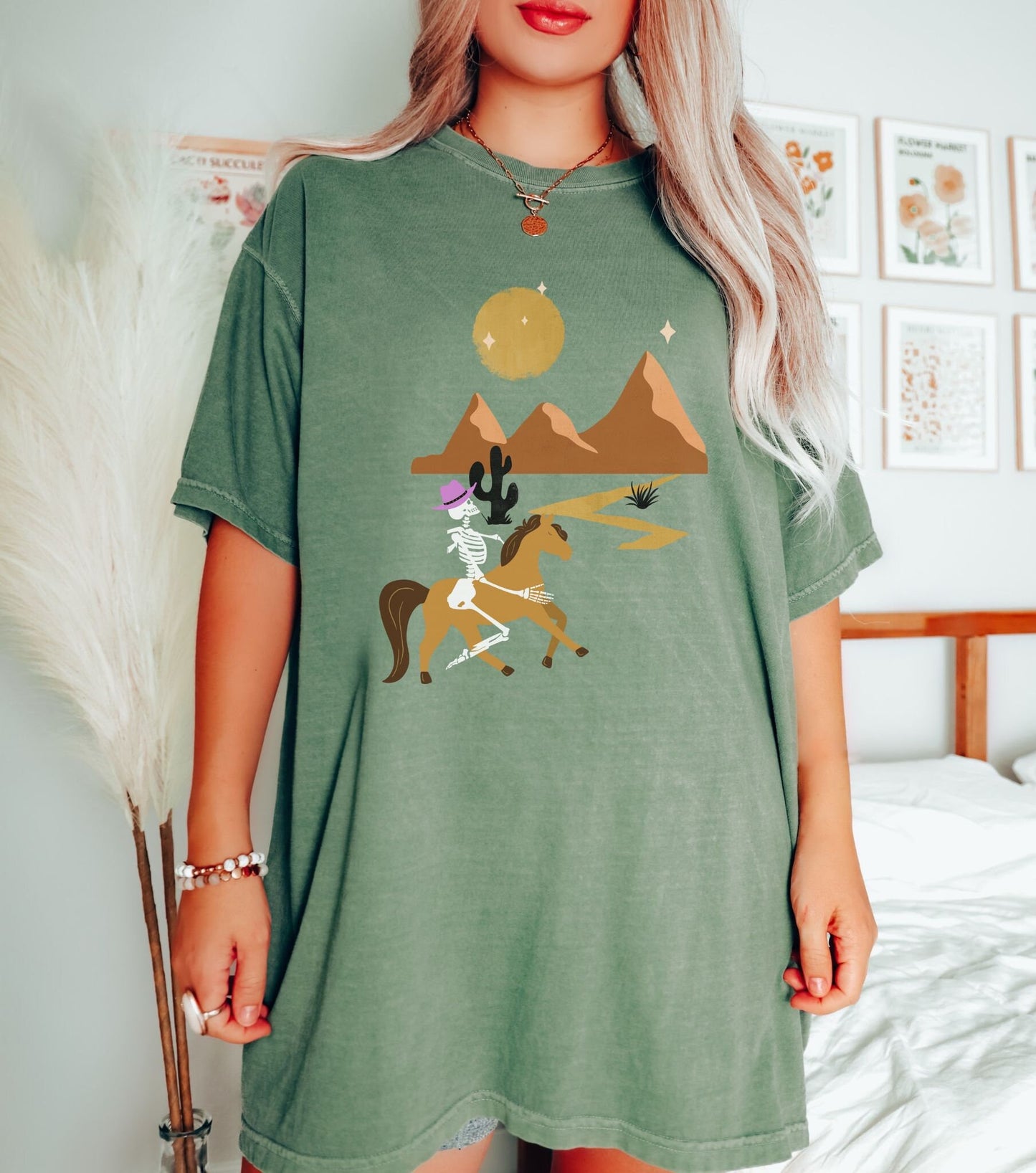 A woman wearing a vintage, moss colored shirt with a skeleton cowgirl wearing a pink cowboy hat riding a brown horse through the eset at night, cacti and three mountains in the background with a full moon and stars in the sky.