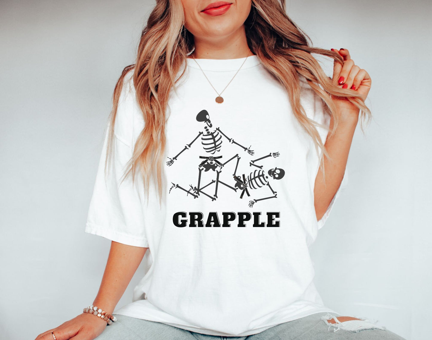 A woman wearing a cute white colored shirt with black text Grapple and two skeletons wearing black belts grappling on the ground.