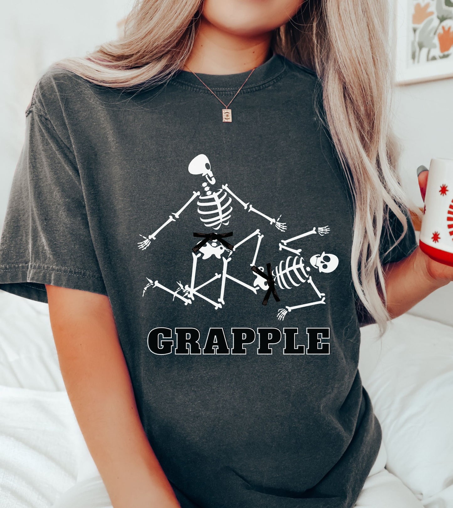 A woman wearing a cute pepper colored shirt with black text Grapple and two skeletons wearing black belts grappling on the ground.