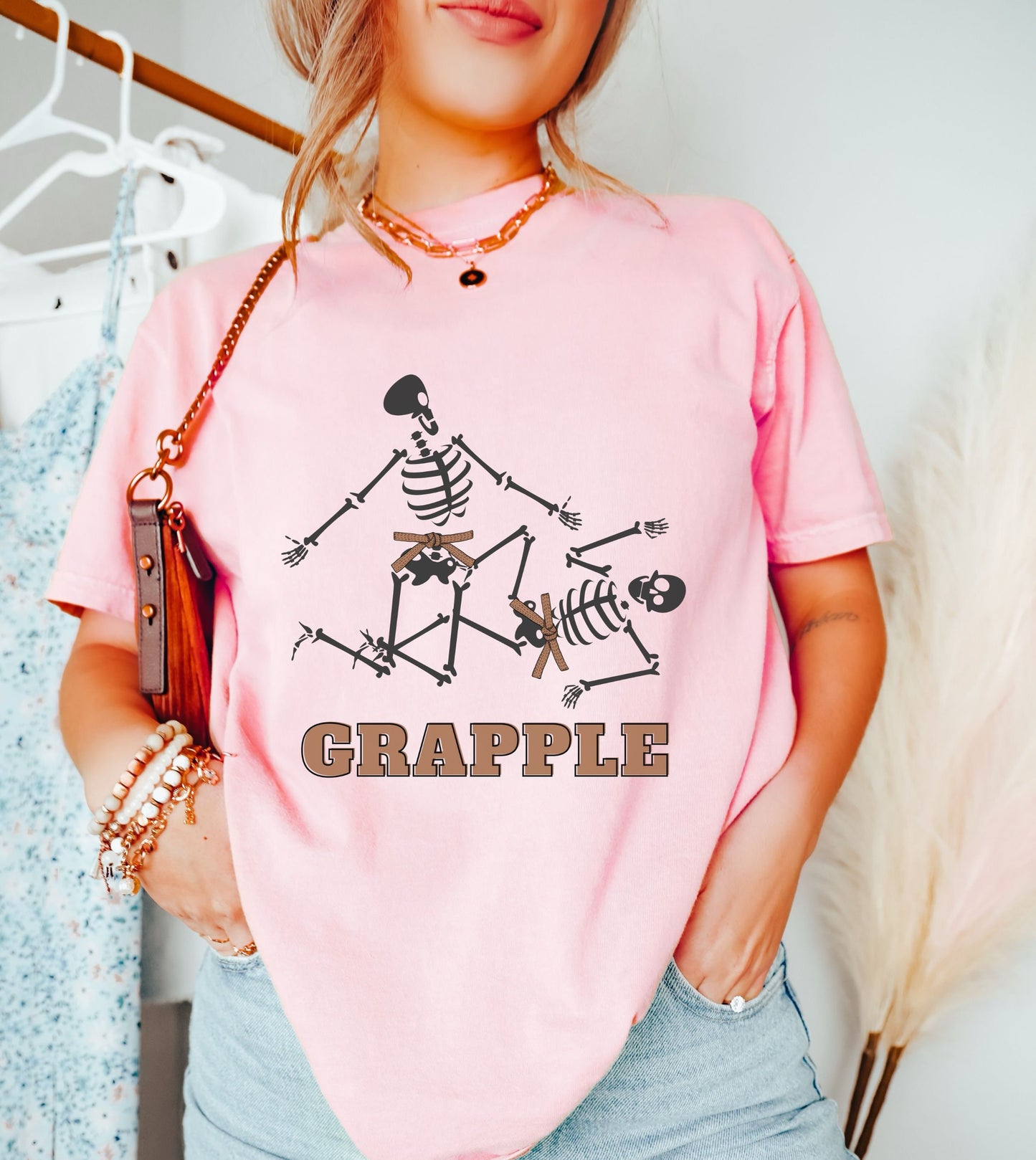 A woman wearing a cute light pink colored shirt with brown text Grapple and two skeletons wearing brown belts grappling on the ground.