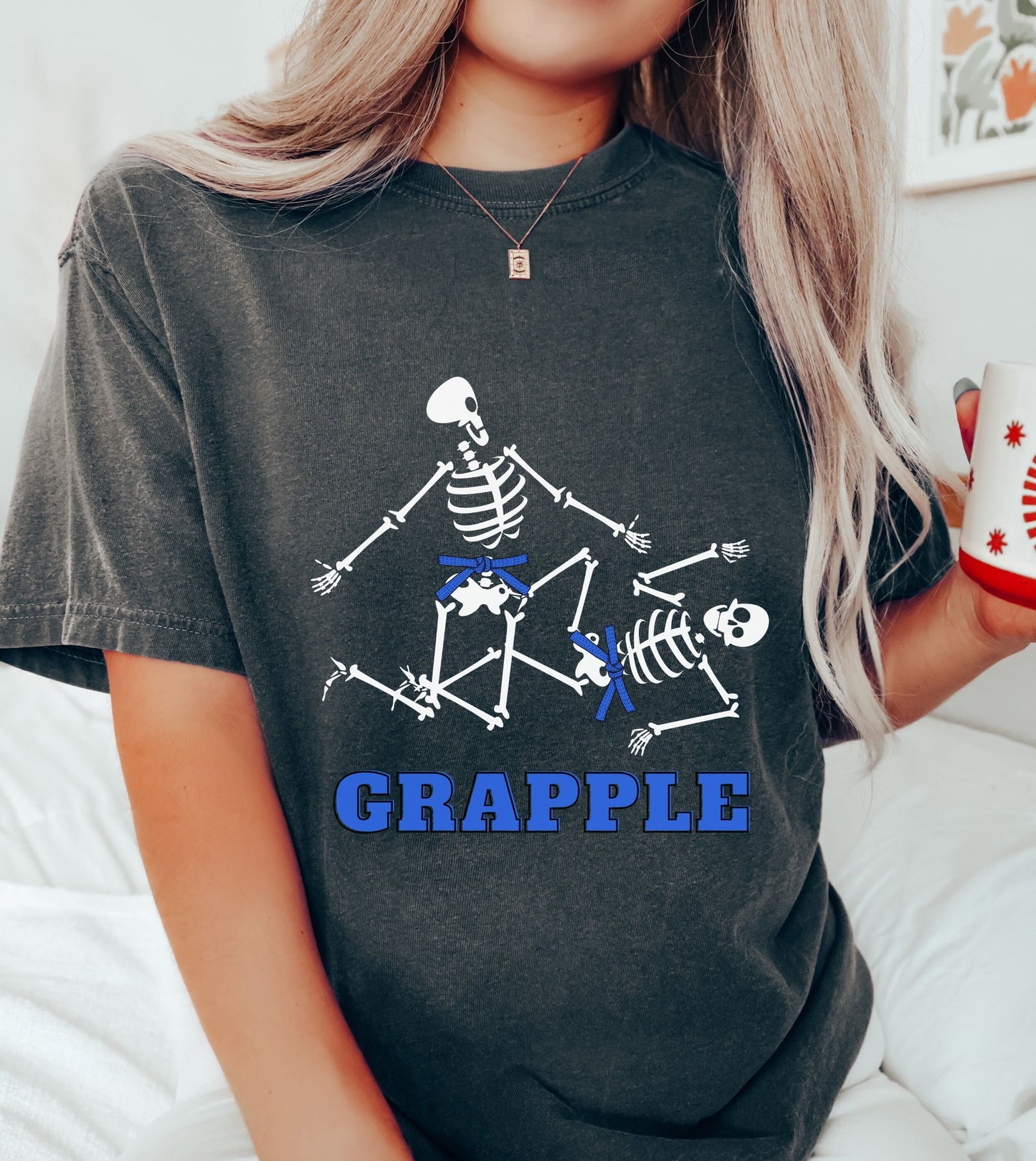 A woman wearing a cute pepper colored shirt with blue text Grapple and two skeletons wearing blue belts grappling on the ground.