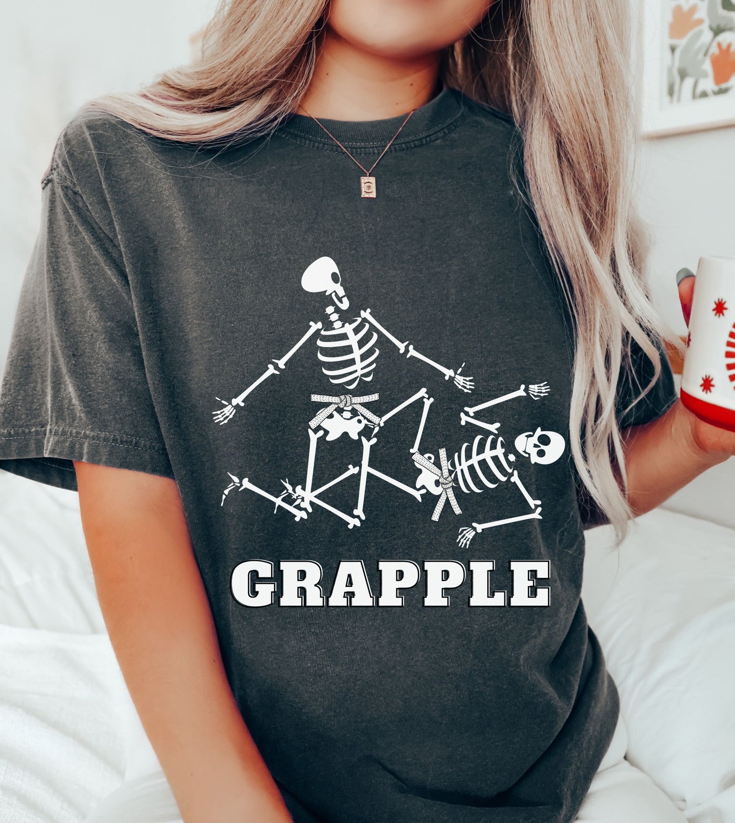 A woman wearing a cute pepper colored shirt with the text Grapple and two skeletons wearing white belts grappling on the ground.