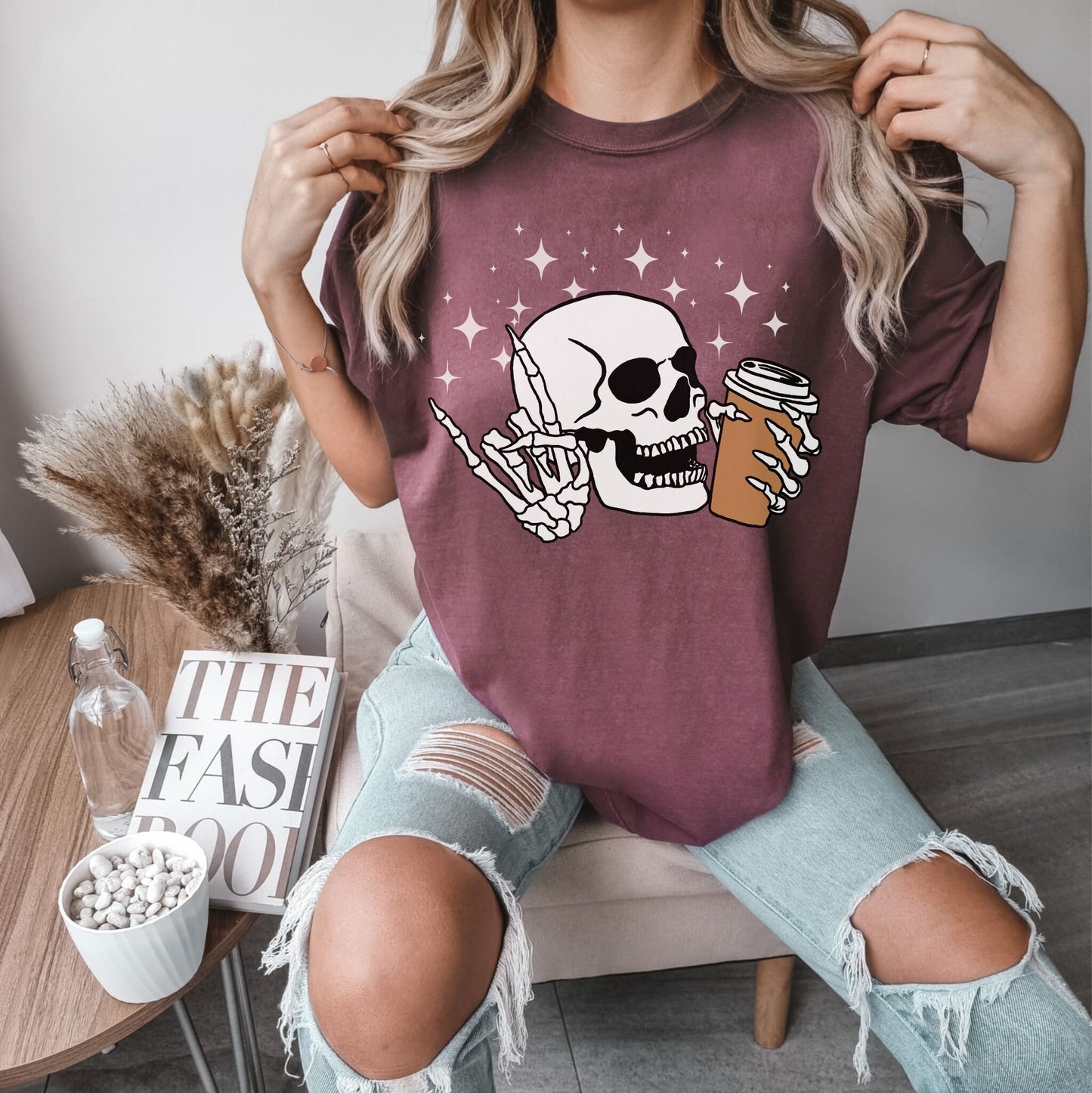 A woman wearing a cute vineyard colored shirt, a skeleton skull holding a coffee in the left hand and holding up the rock on hand sign with the right hand with twinkly stars in the background.