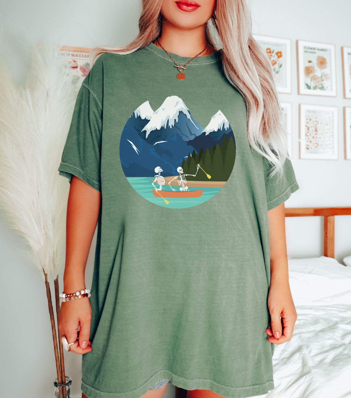 A woman wearing a cute, vintage moss colored shirt with a picture of a skeleton couple rowing a canoe with yellow paddles through a river with snow-capped mountains in the background.