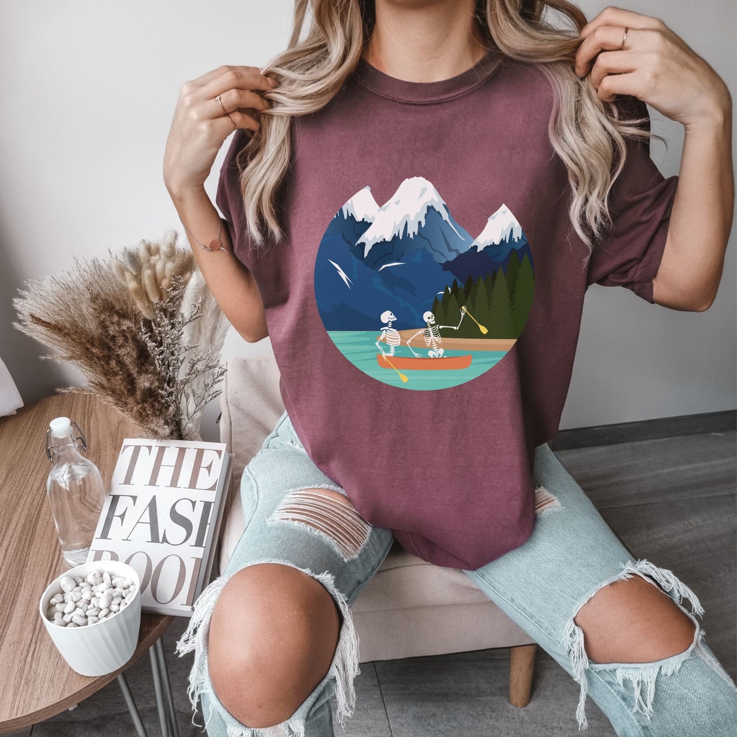 A woman wearing a cute, vintage vineyard colored shirt with a picture of a skeleton couple rowing a canoe with yellow paddles through a river with snow-capped mountains in the background.