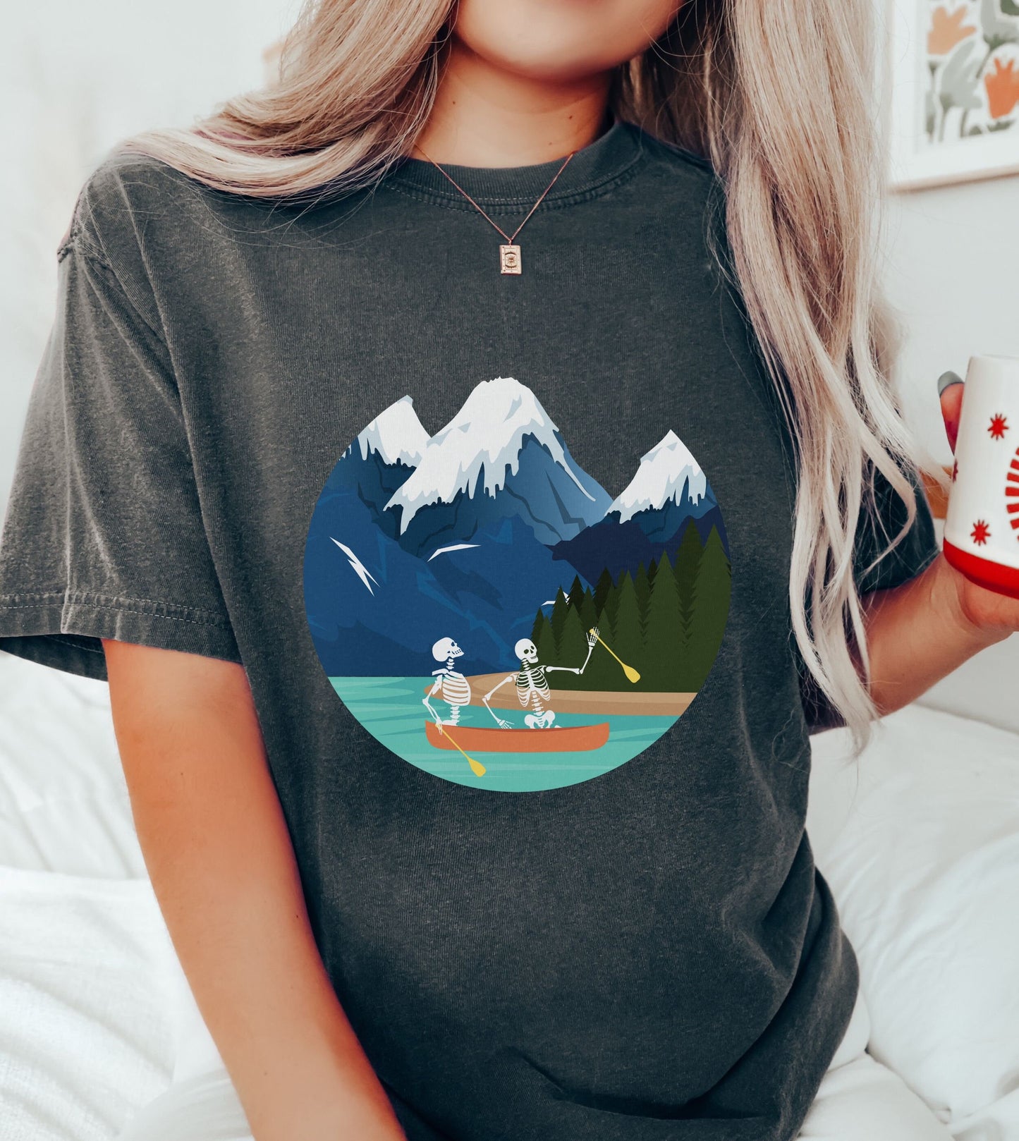 A woman wearing a cute, vintage pepper colored shirt with a picture of a skeleton couple rowing a canoe with yellow paddles through a river with snow-capped mountains in the background.