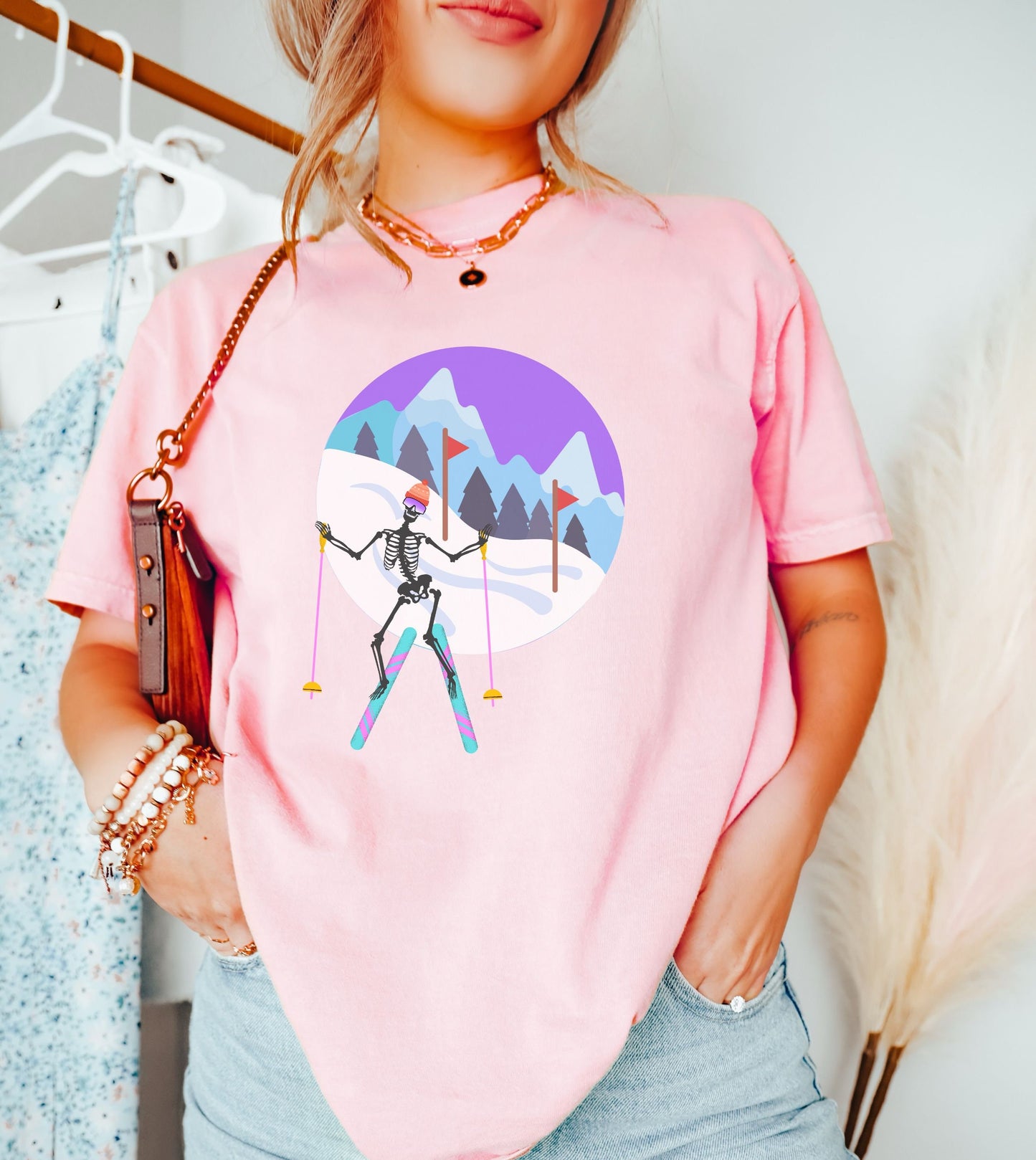 A woman wearing a vintage, blossom colored shirt with a female skeleton wearing snow goggles and a beanie skiing with pink and turquoise skis and pink ski poles, red slalom flags and trees and mountains in the background in front of a purple skyline.