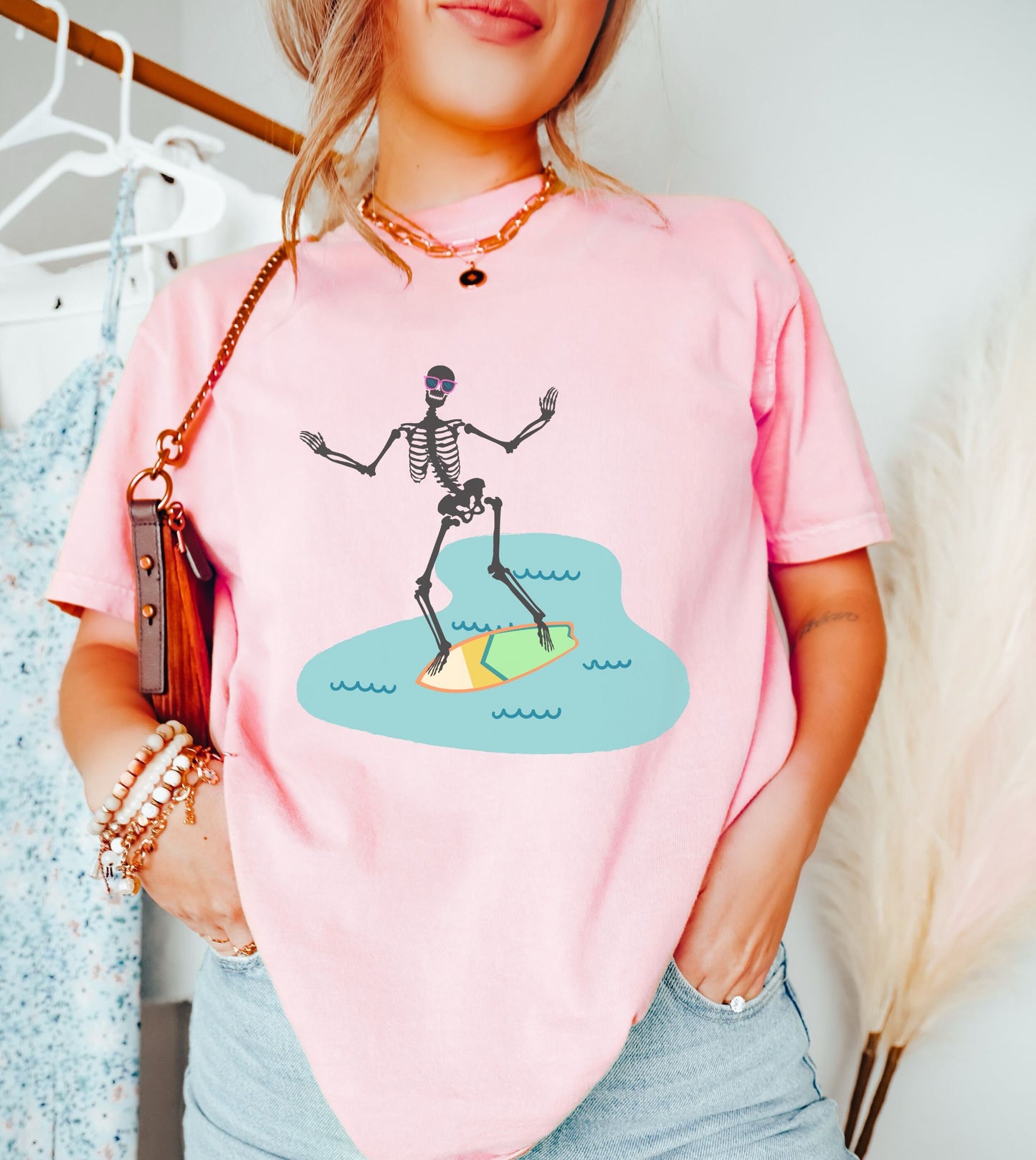 A woman wearing a cute, vintage blossom colored shirt with a skeleton wearing pink sunglasses and balancing on a green and yellow surfboard or wakeboard in the water.