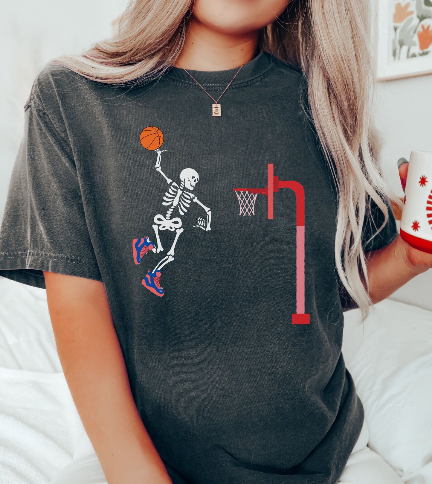A woman wearing a cute pepper colored shirt of a skeleton with colorful basketball shoes in mid air dunking a basketball on a goal.
