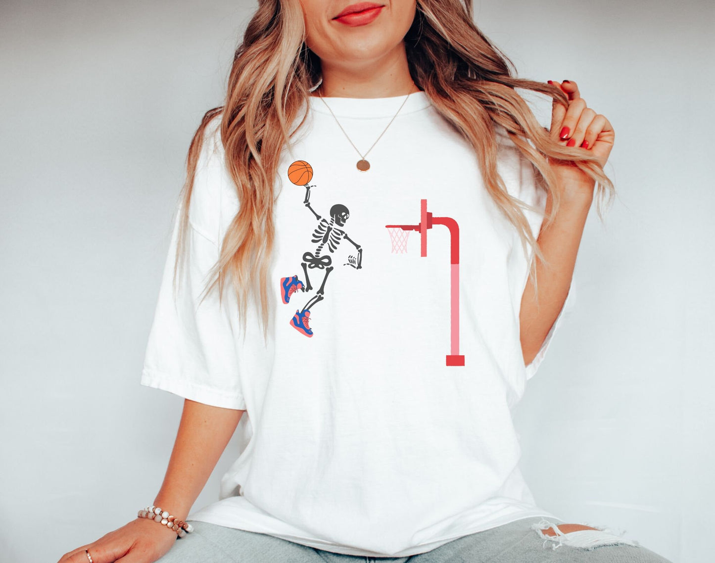 A woman wearing a cute white colored shirt of a skeleton with colorful basketball shoes in mid air dunking a basketball on a goal.