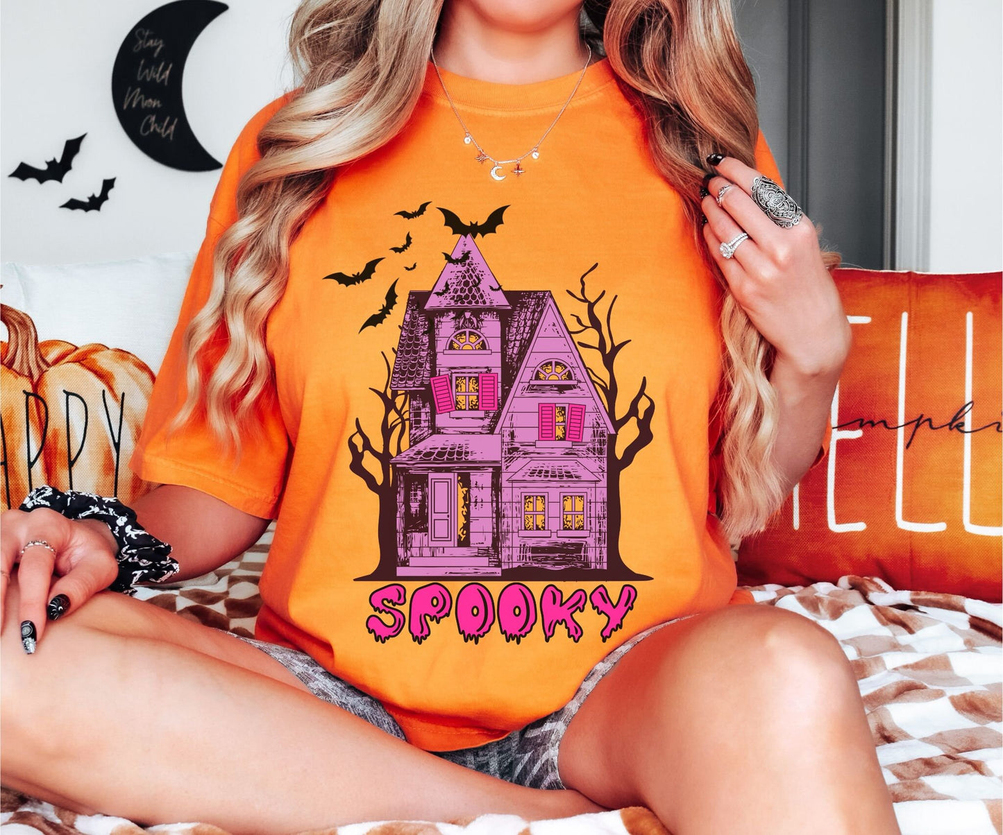 A woman wearing a cute, vintage burnt orange colored Comfort Colors T-shirt with the text Spooky in purple across the bottom. Above is a Purple and pink haunted house with dark trees in the background with black bats flying above.