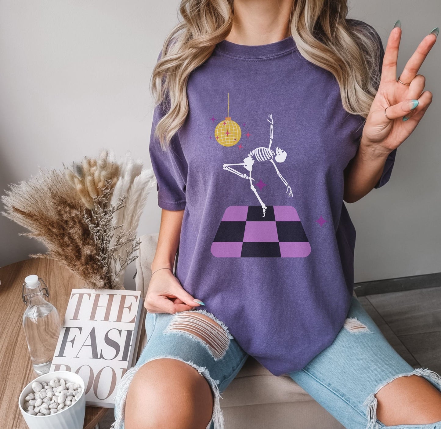 A woman wearing a cute purple colored shirt with a female skeleton dancing on a black and purple dance floor under a yellow, spinning disco ball.