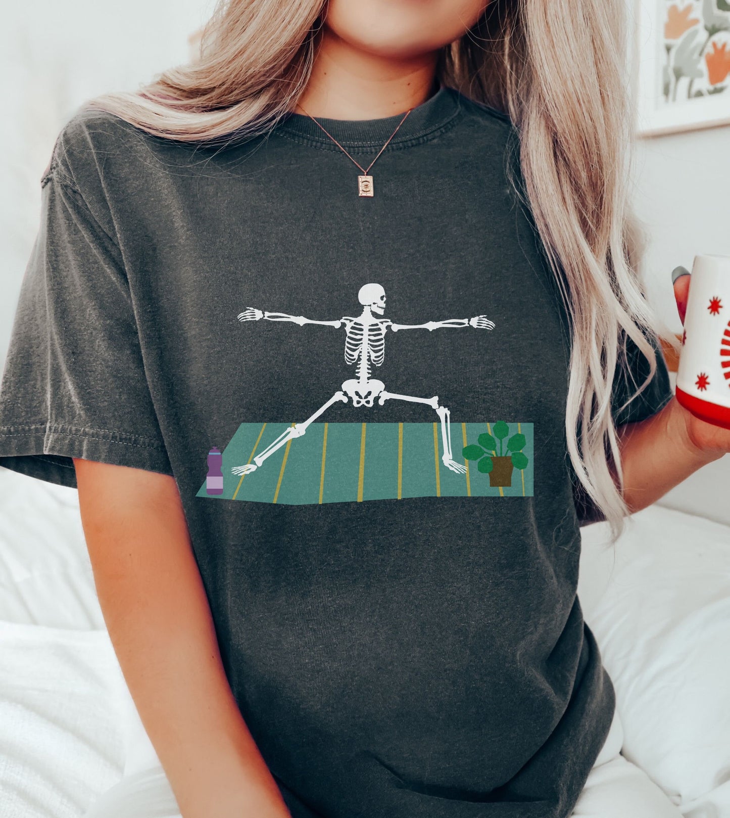 A woman wearing a vintage, pepper colored shirt with a white skeleton performing a yoga pose on a green and yellow towel with a purple water bottle and potted plant on the towel by the yogi’s feet.