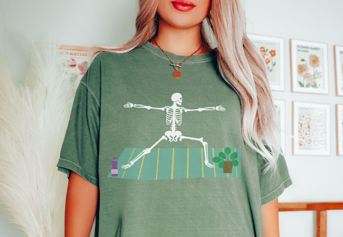 A woman wearing a vintage, moss colored shirt with a white skeleton performing a yoga pose on a green and yellow towel with a purple water bottle and potted plant on the towel by the yogi’s feet.