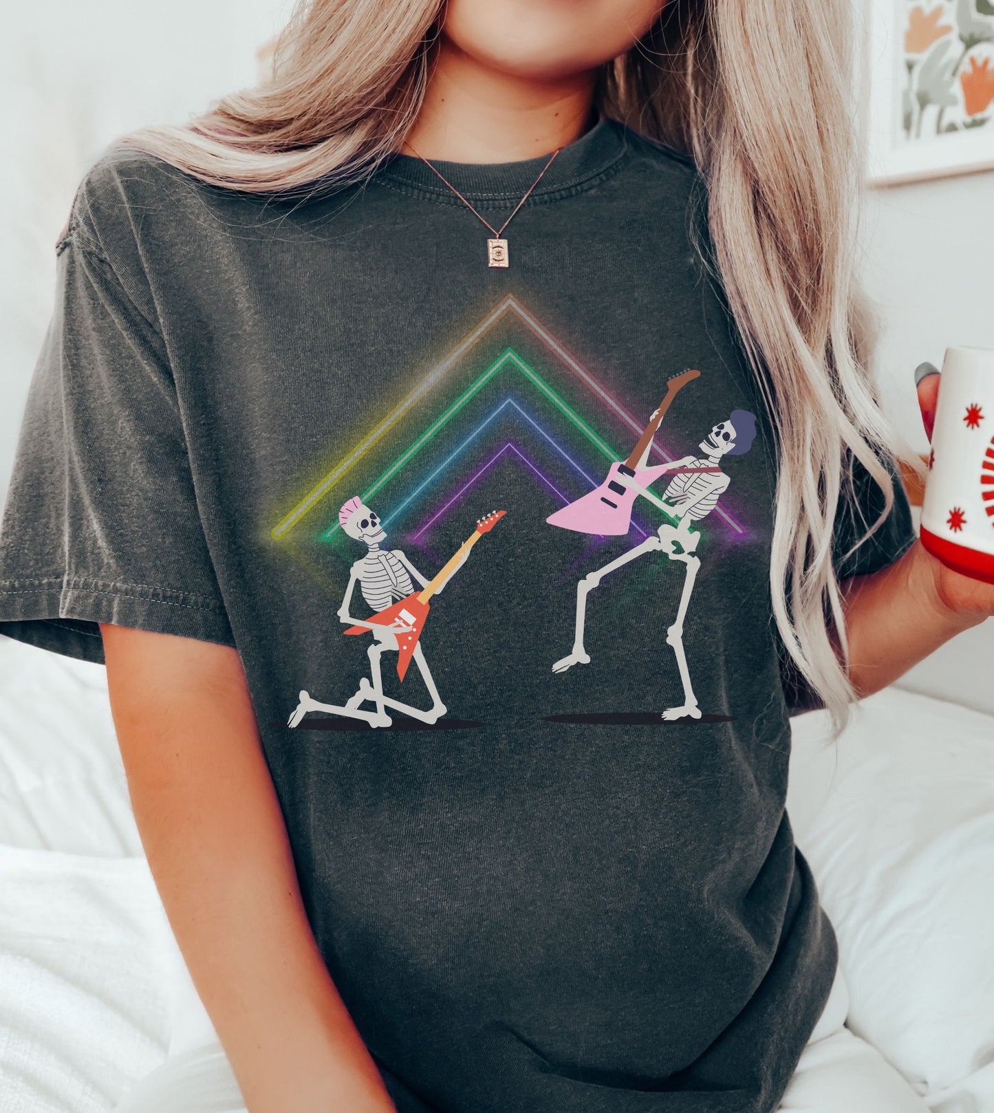 A woman wearing a cute pepper colored shirt with two skeletons rocking fun hairstyles jamming out on electric guitars in front of a neon lights setup. One skeleton is on its knees and the other has one leg in the air, both rocking out music.