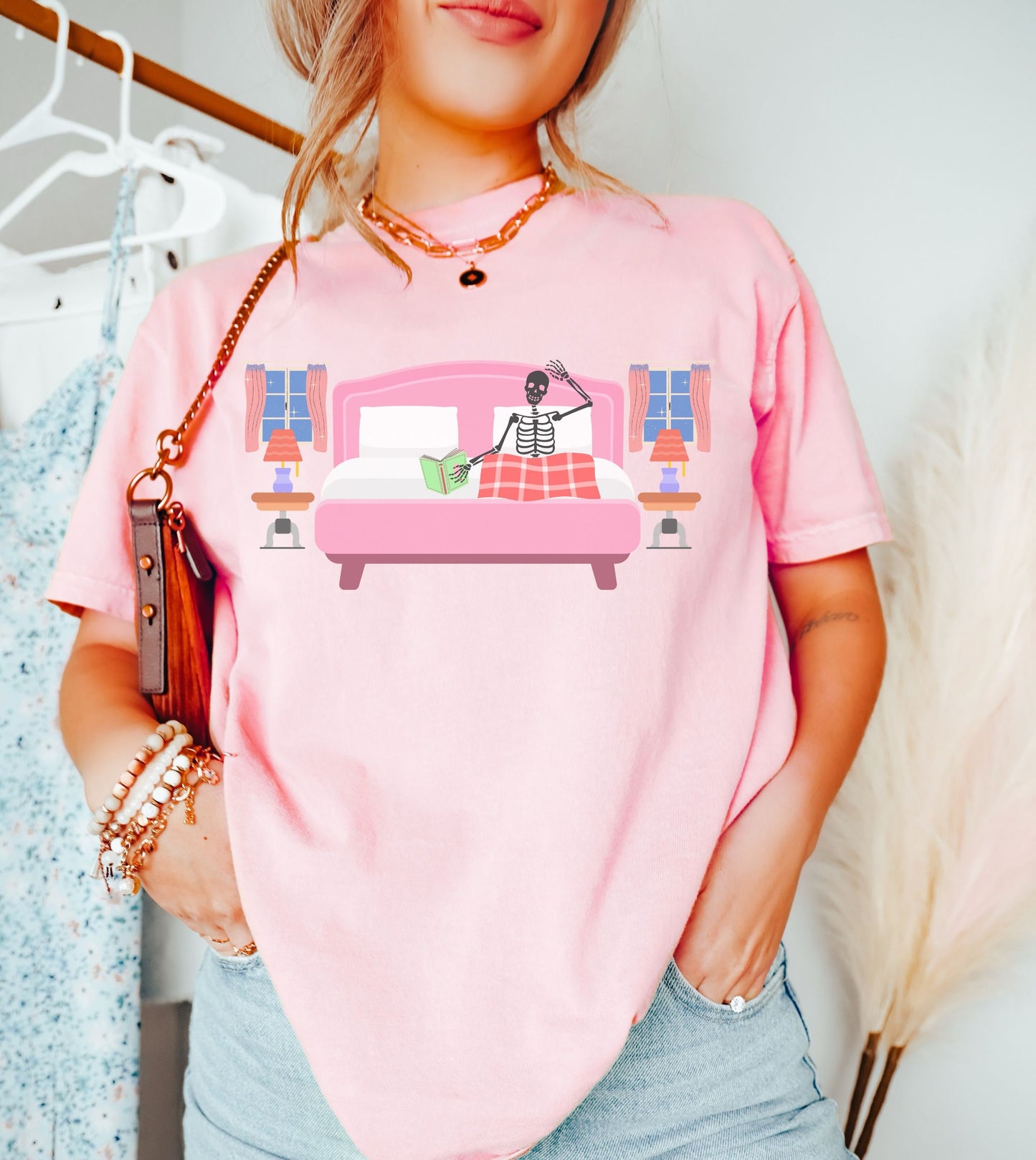 A woman wearing a cute blossom colored shirt with a skeleton lying in a pink bed covered with a red blanket reading a green book, two night tables and windows on each side of the bed.