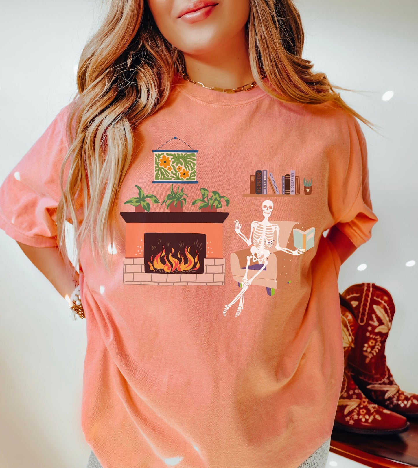 A woman wearing a cute terracotta colored shirt with a picture of a skeleton sitting in a cushy chair reading a book under a bookshelf, a fireplace with a roaring fire to the right of the chair with plants on top of the hearth.