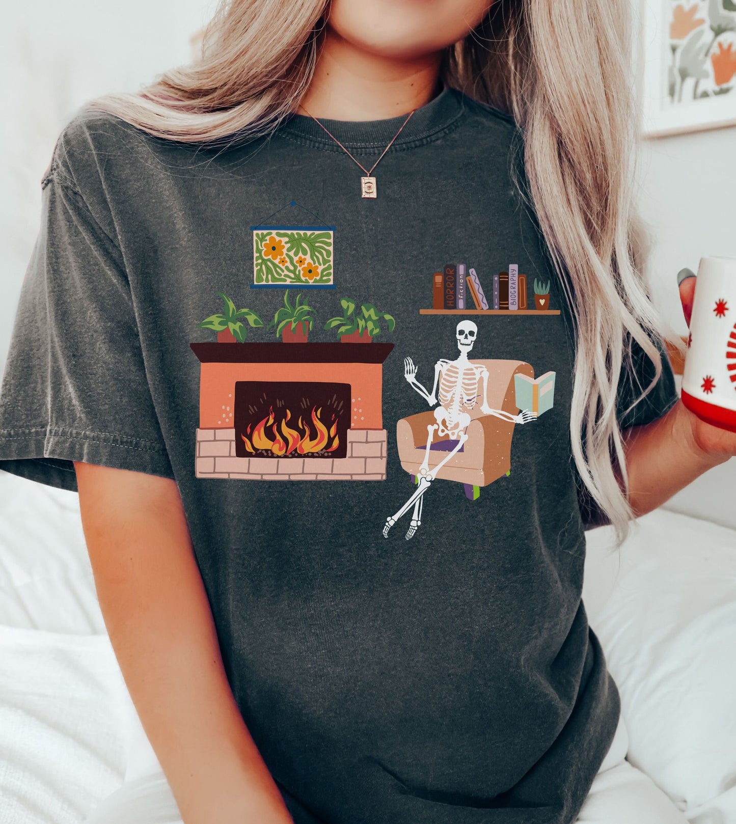 A woman wearing a cute black colored shirt with a picture of a skeleton sitting in a cushy chair reading a book under a bookshelf, a fireplace with a roaring fire to the right of the chair with plants on top of the hearth.