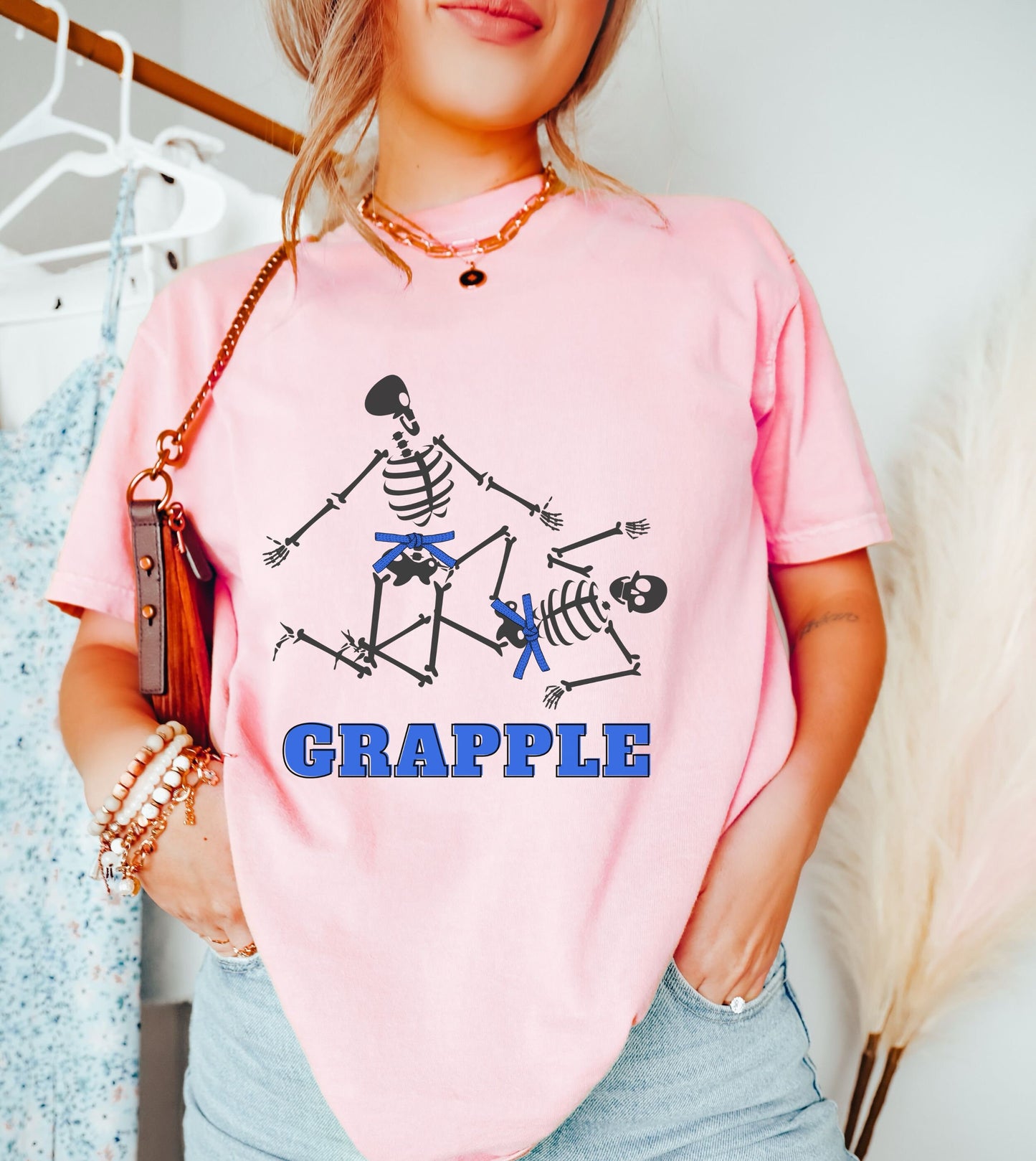 A woman wearing a cute light pink colored shirt with blue text Grapple and two skeletons wearing blue belts grappling on the ground.