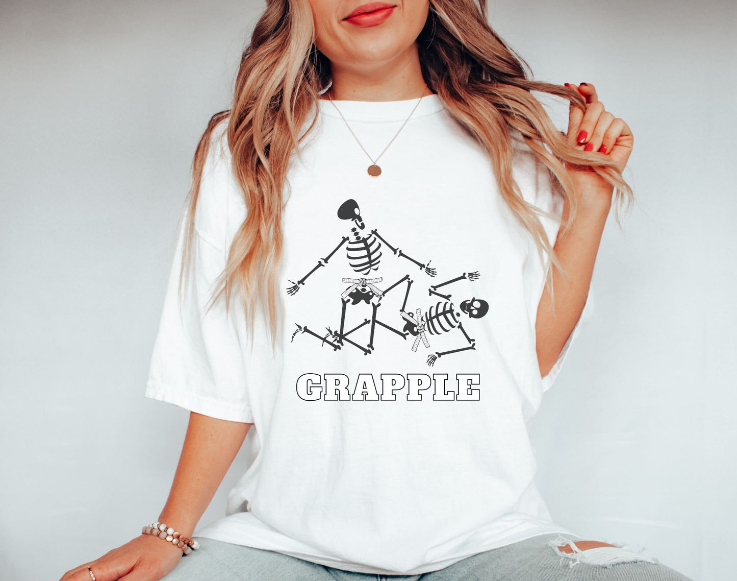 A woman wearing a cute white colored shirt with the text Grapple and two skeletons wearing white belts grappling on the ground.