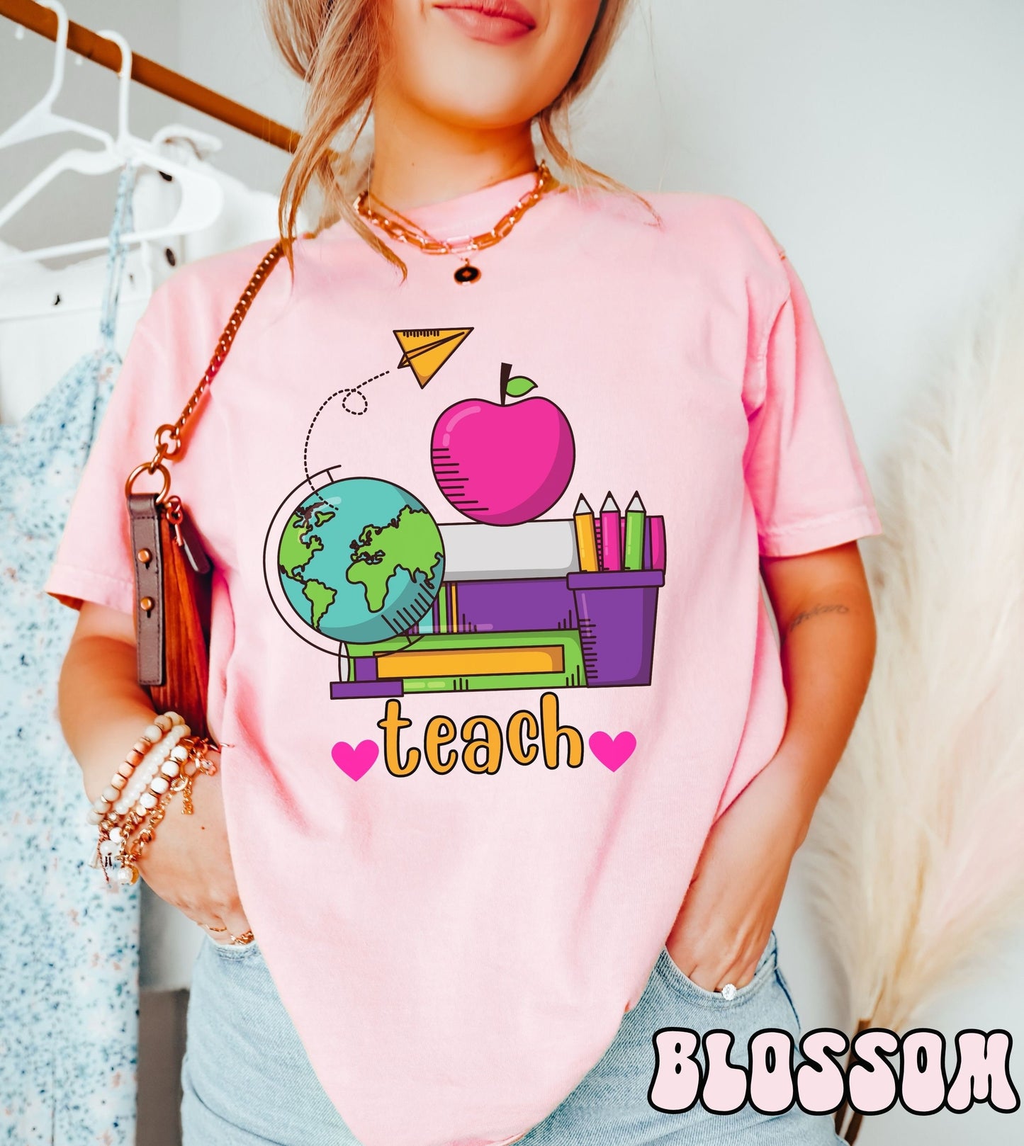 A woman wearing a vintage, blossom colored shirt with the word teach in between two pink hearts, and above are a stack of green, purple, and white books, a pink apple and a colorful pencil holder, a globe, and a yellow paper airplane flying above.