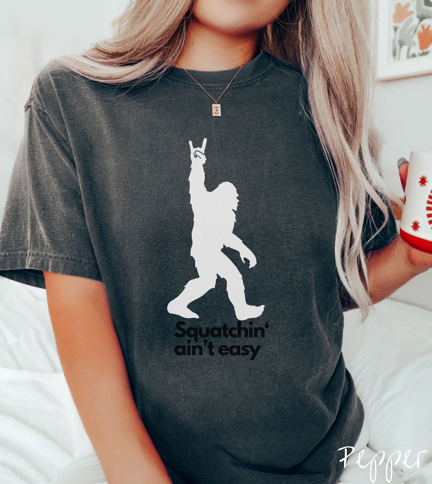 A woman wearing a cute pepper colored shirt with text saying Squatchin aint easy and a silhouette of Bigfoot holding up the rock on hand sign.