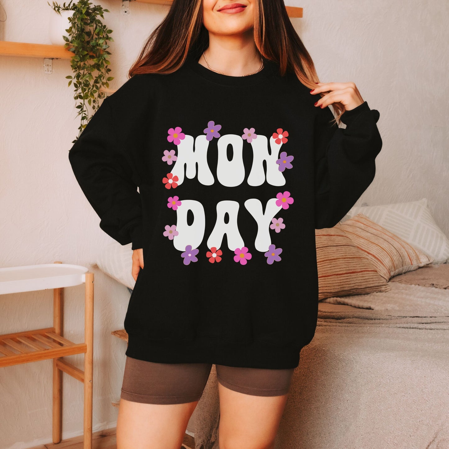 A woman wearing a vintage, black colored sweatshirt with the text centered on the front MON then DAY below it, with pink, purple, white, and orange flowers scattered amongst the letters.