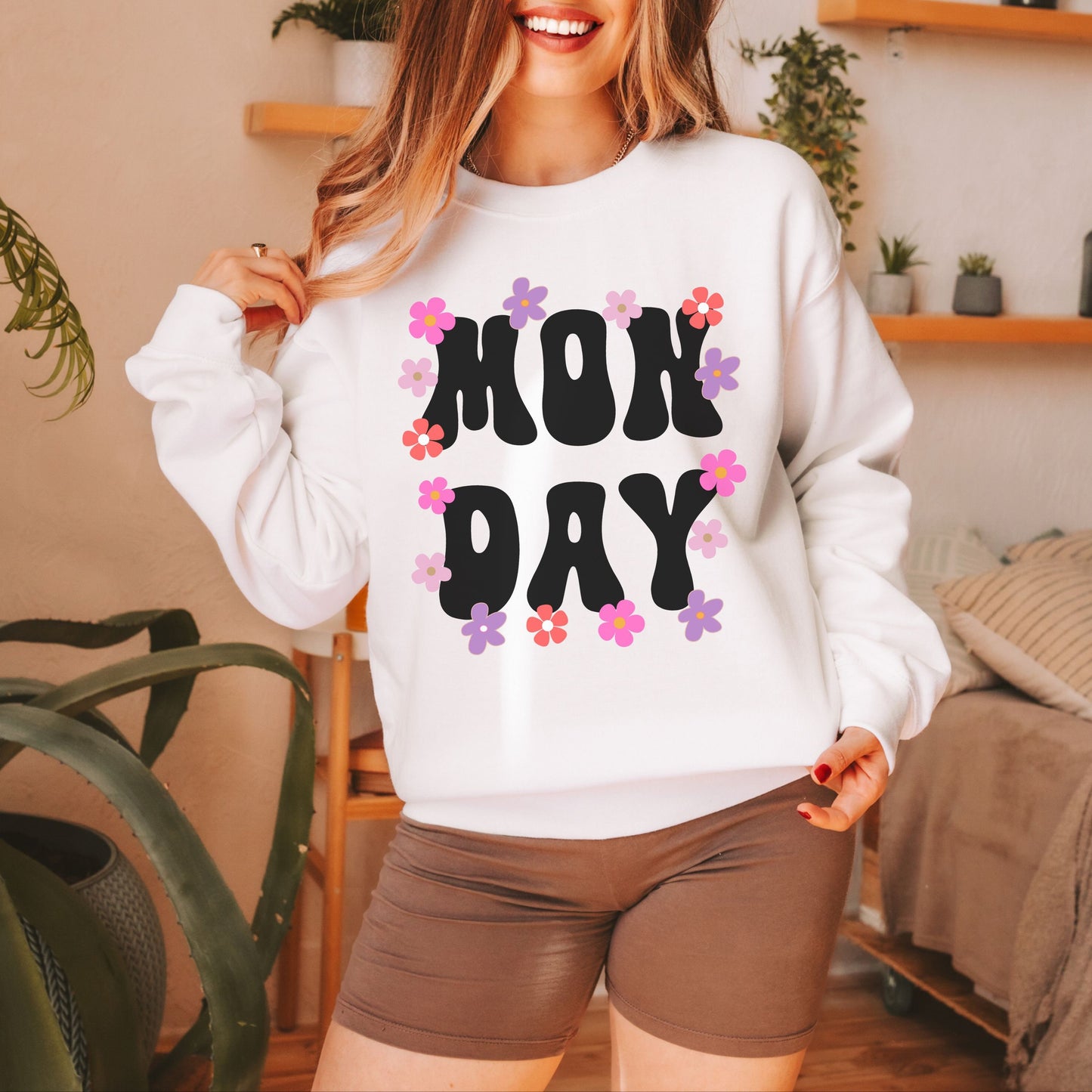 A woman wearing a vintage, white colored sweatshirt with the text centered on the front MON then DAY below it, with pink, purple, white, and orange flowers scattered amongst the letters.