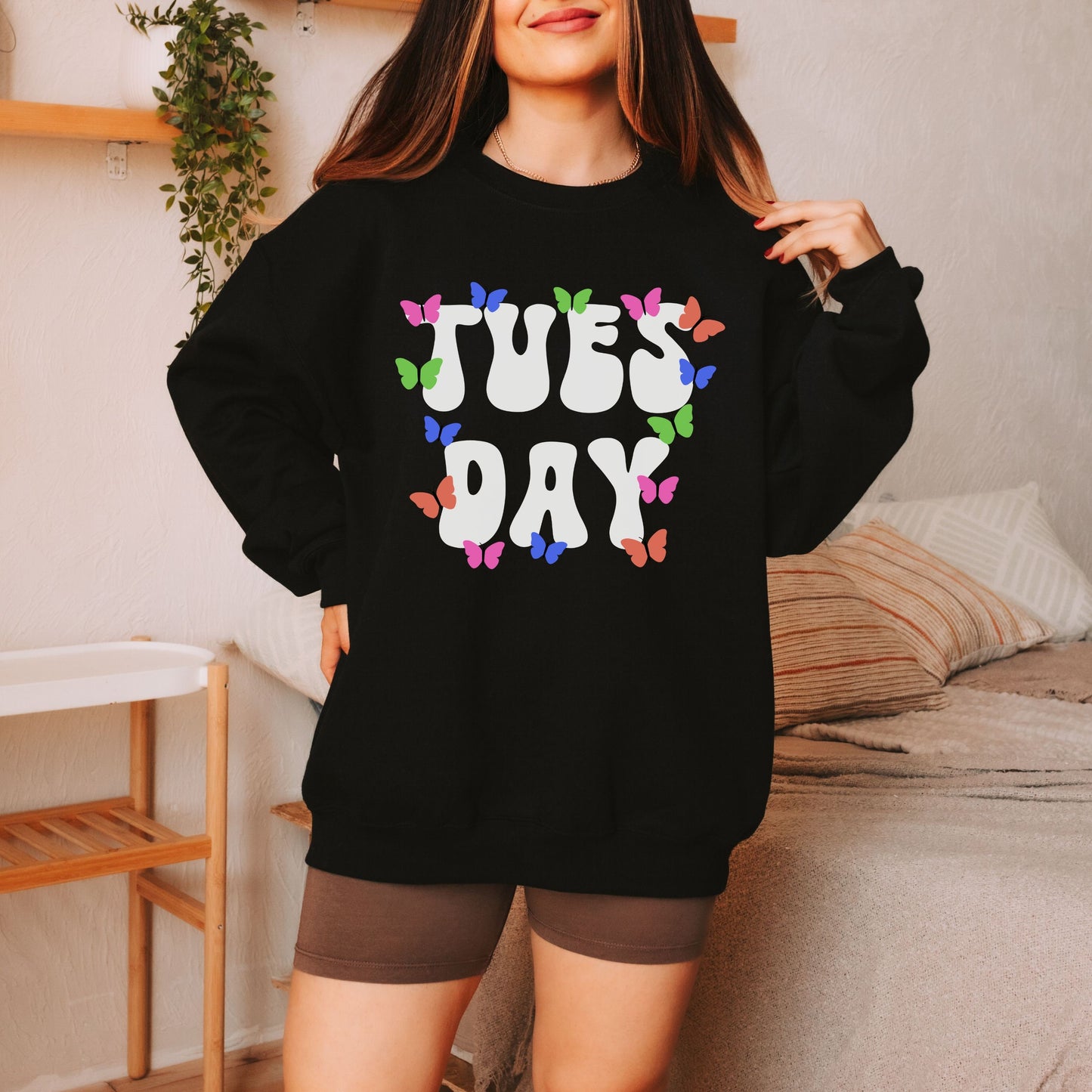 A woman wearing a vintage, black colored sweatshirt with the text centered on the front TUES then DAY below it, with pink, green, blue, and orange butterflies scattered amongst the letters.