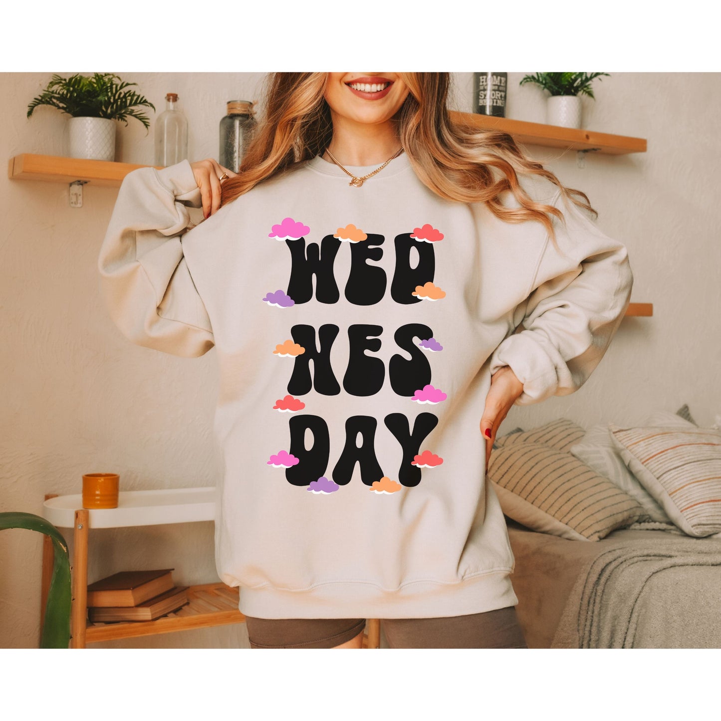 A woman wearing a vintage, sand colored sweatshirt with the text centered on the front WED then below it NES then DAY below that, with pink, red, purple, and orange clouds scattered amongst the letters.