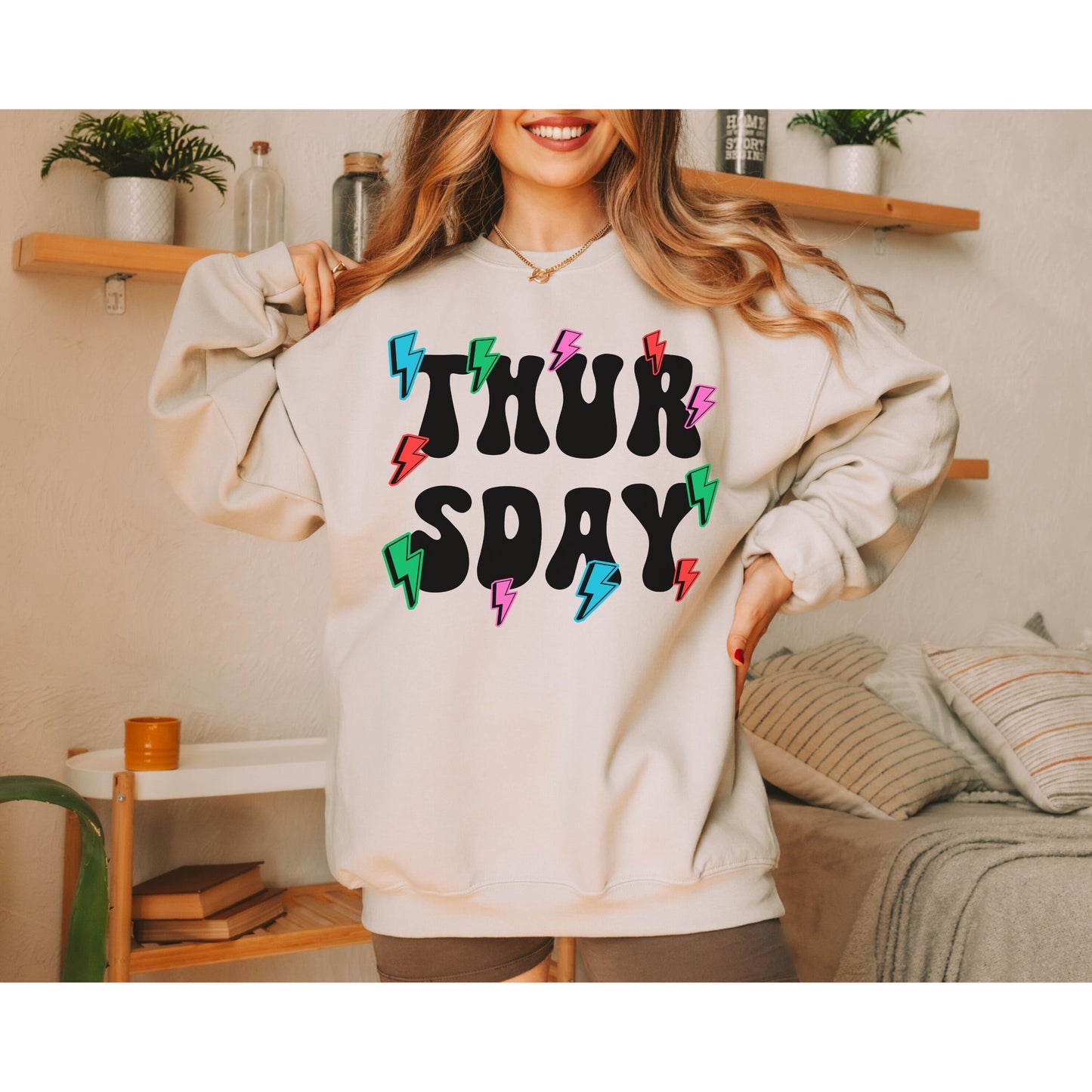 A woman wearing a vintage, sand colored sweatshirt with the text centered on the front THURS then below it DAY, with pink, red, blue, and green lightning bolts scattered amongst the letters.