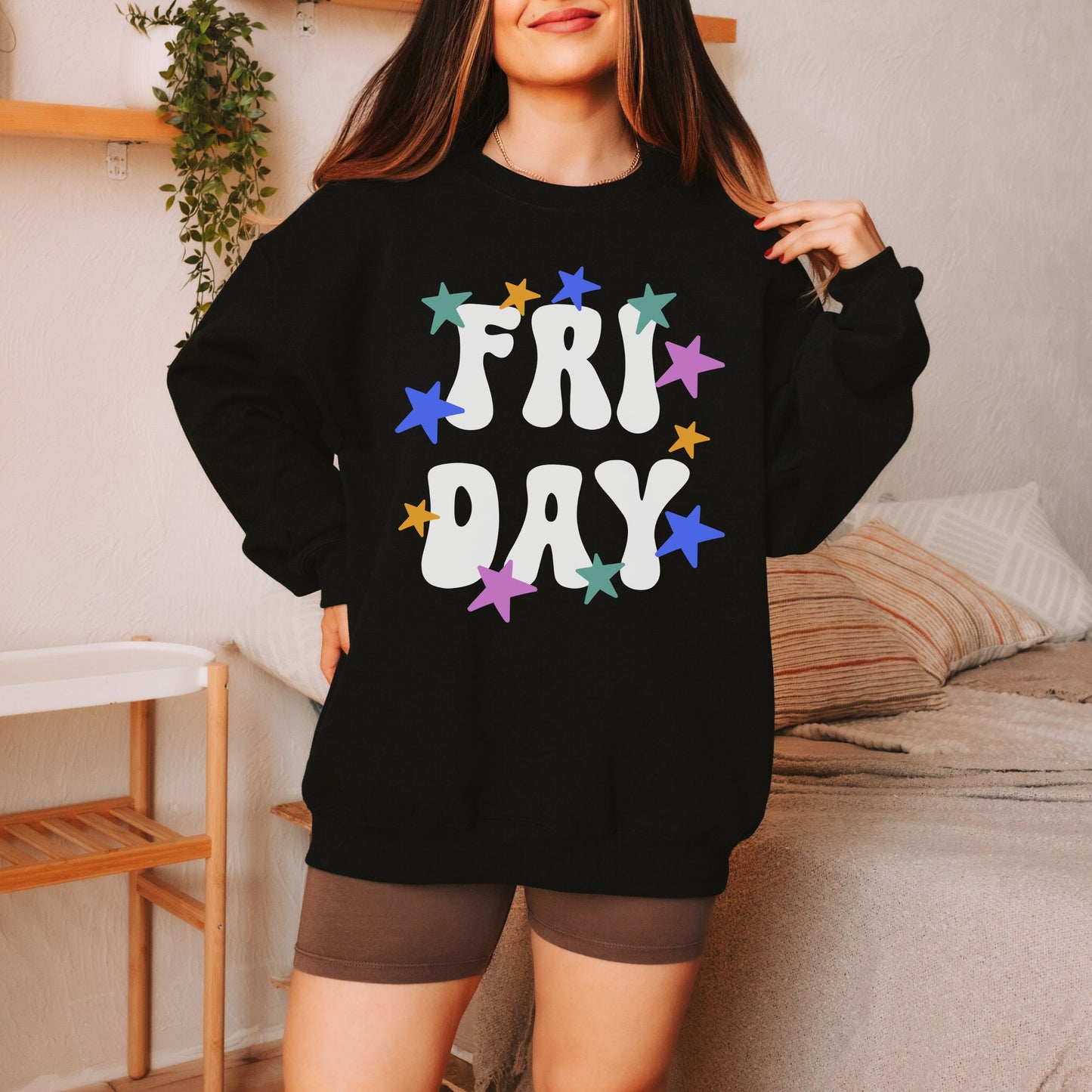 A woman wearing a vintage, black colored sweatshirt with the text centered on the front FRI then below it DAY, with pink, gold, blue, and green stars scattered amongst the letters.