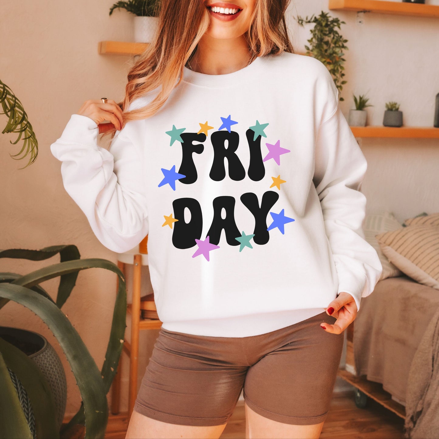 A woman wearing a vintage, white colored sweatshirt with the text centered on the front FRI then below it DAY, with pink, gold, blue, and green stars scattered amongst the letters.