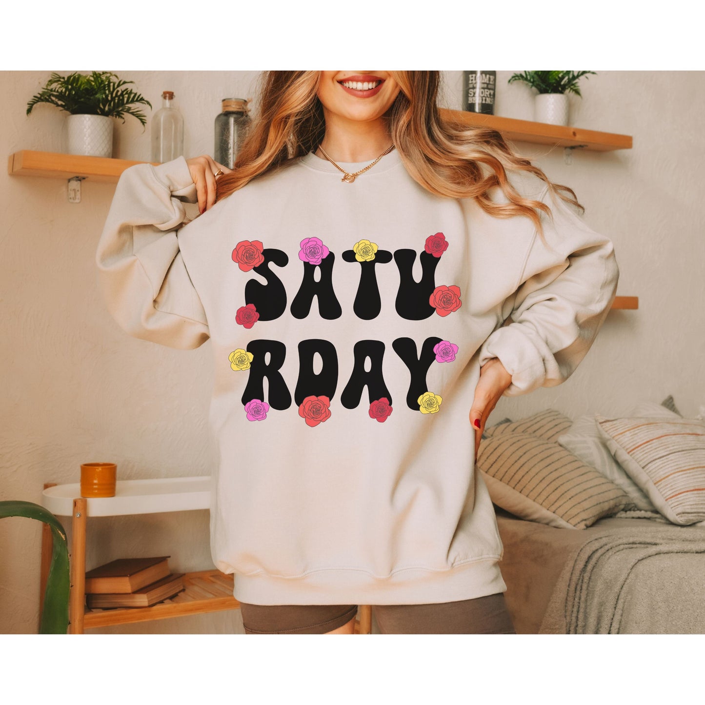 A woman wearing a vintage, sand colored sweatshirt with the text centered on the front SATU then below it RDAY, with pink, red, and yellow roses scattered amongst the letters.