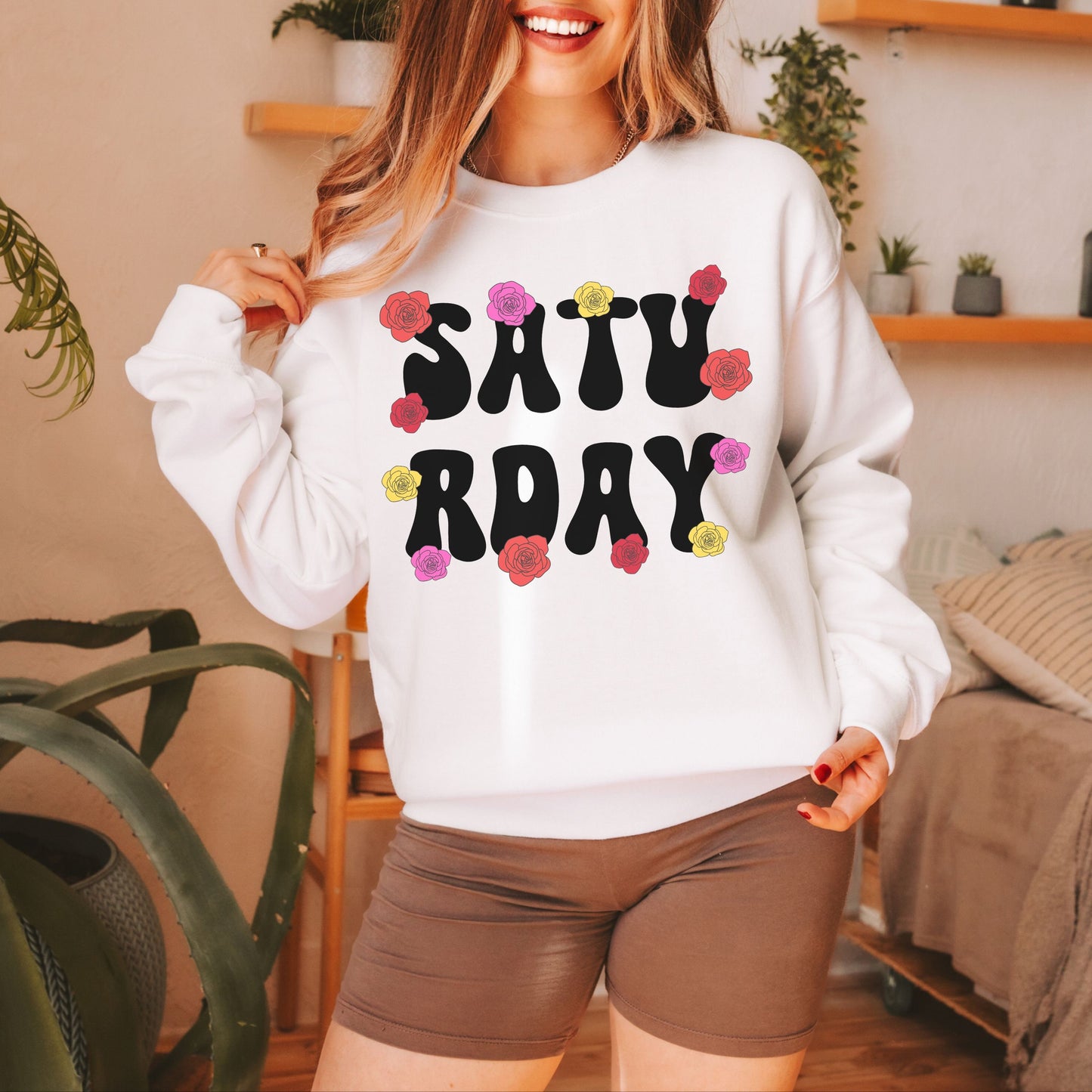 A woman wearing a vintage, white colored sweatshirt with the text centered on the front SATU then below it RDAY, with pink, red, and yellow roses scattered amongst the letters.
