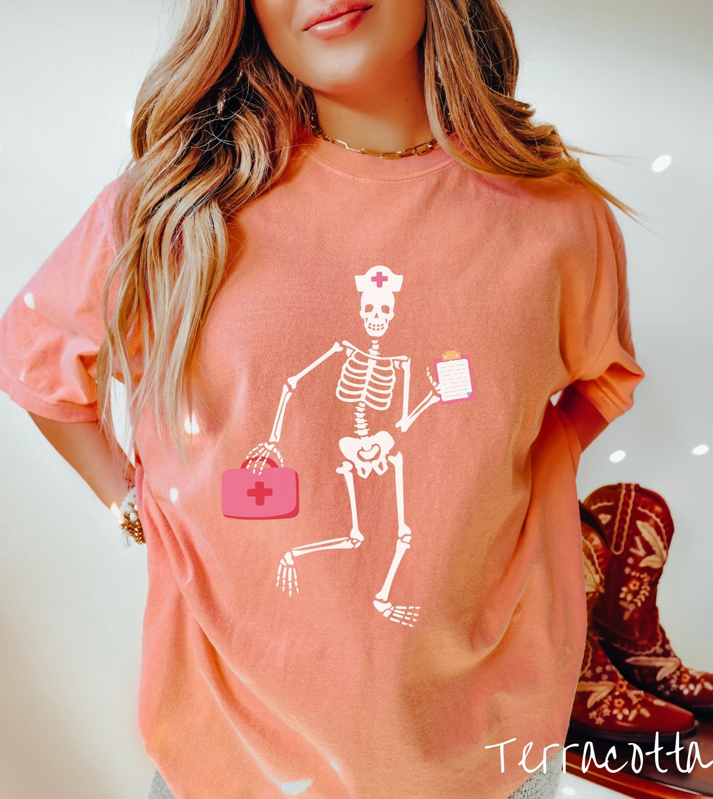 A woman wearing a cute terracotta colored shirt with a female skeleton dancing wearing a nurse’s hat and carrying a pink medical bag and a pink clipboard.